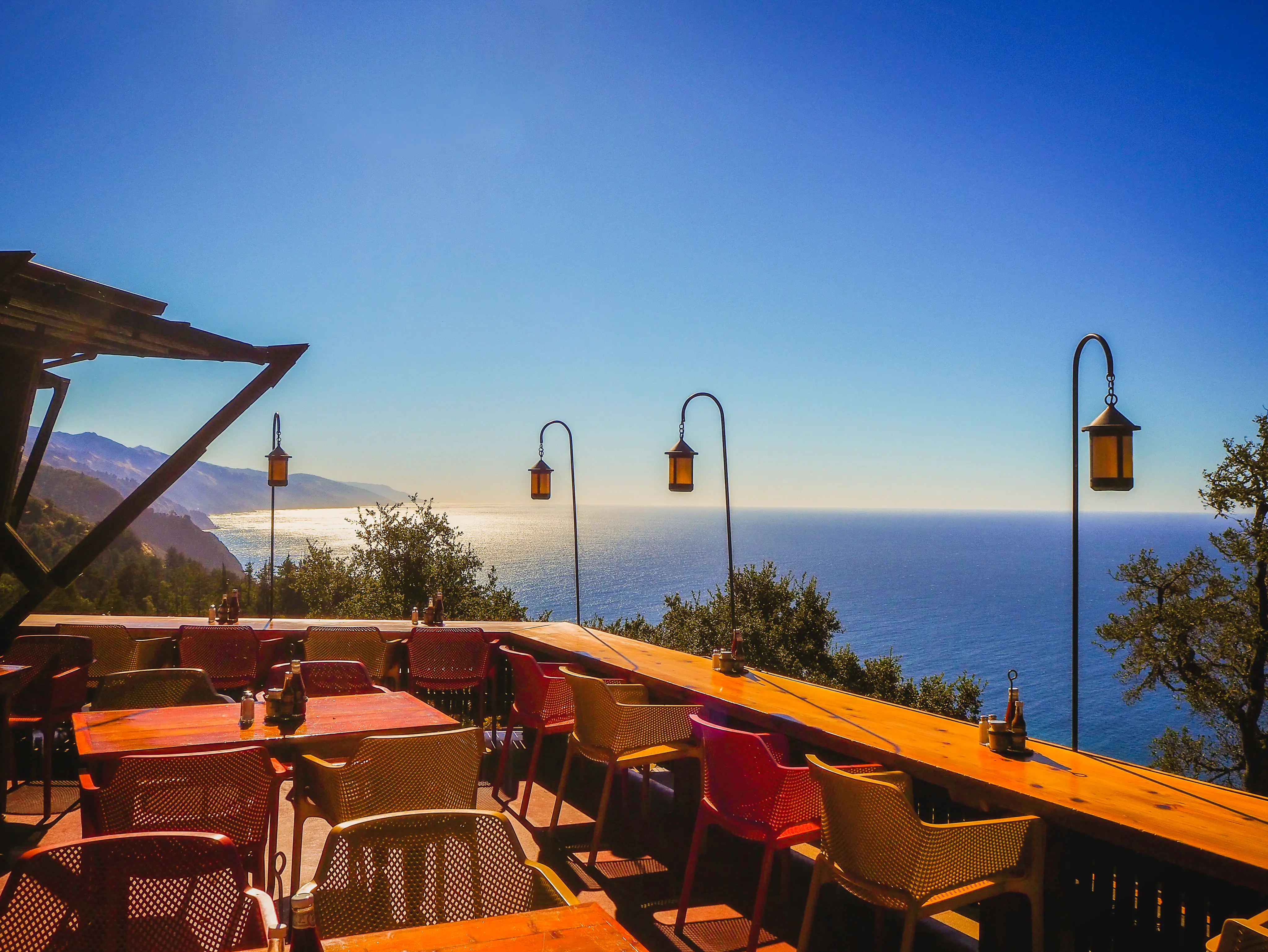 Outdoor terrace dining area with tables, chairs, hanging lanterns, and a panoramic ocean coastline view.