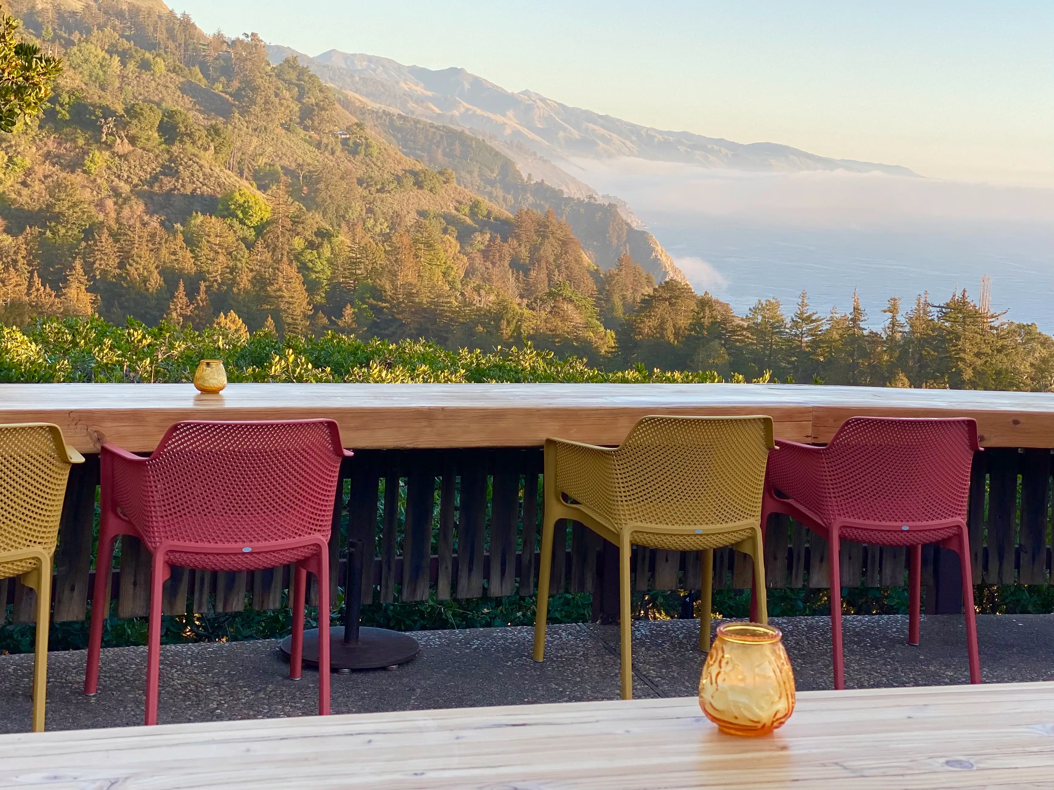 Outdoor terrace seating overlooks forested coastal mountains and ocean fog in warm sunlight.