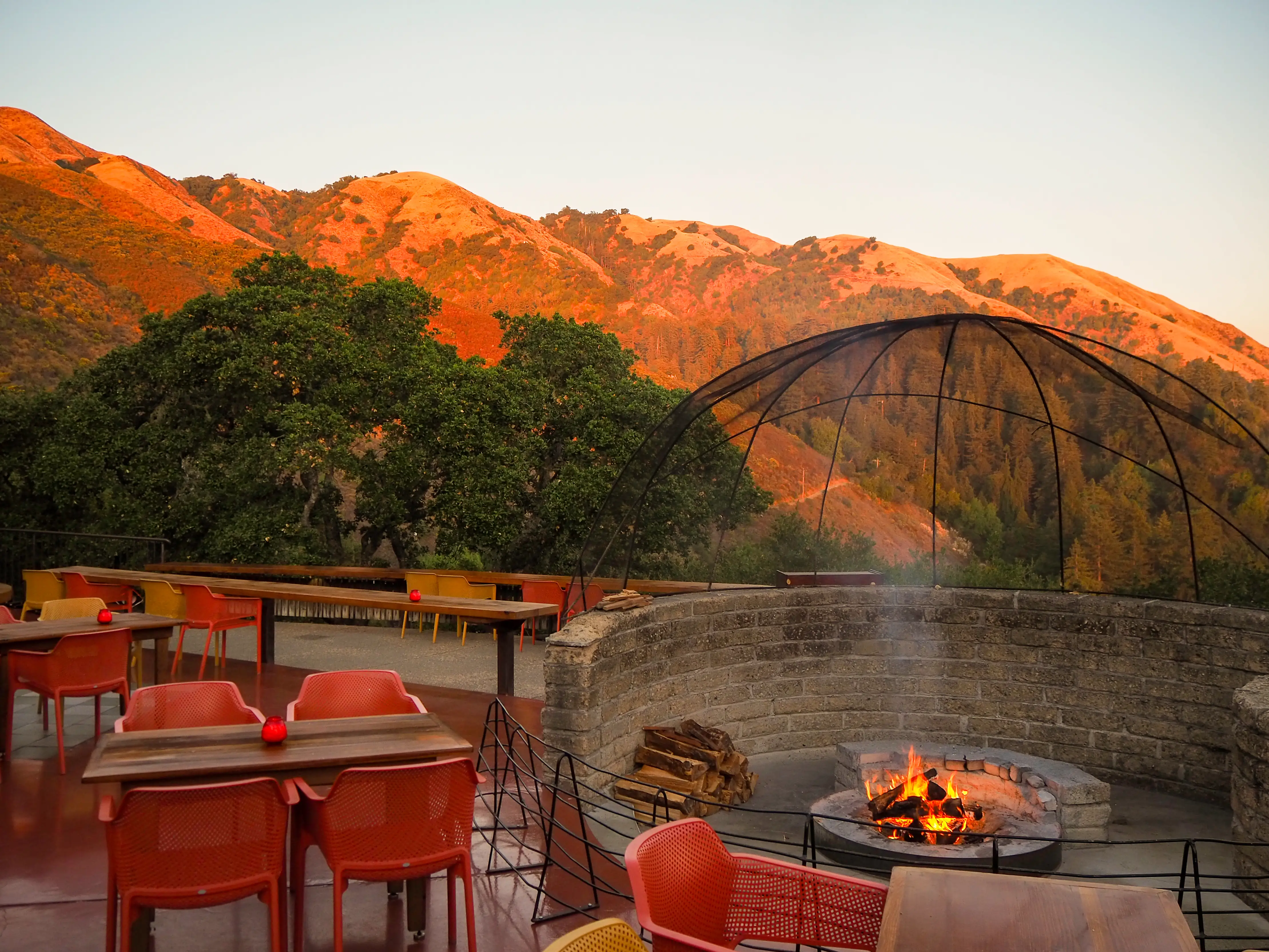 Outdoor terrace dining area with red chairs, a fire pit, trees, and sunlit hills in the background.