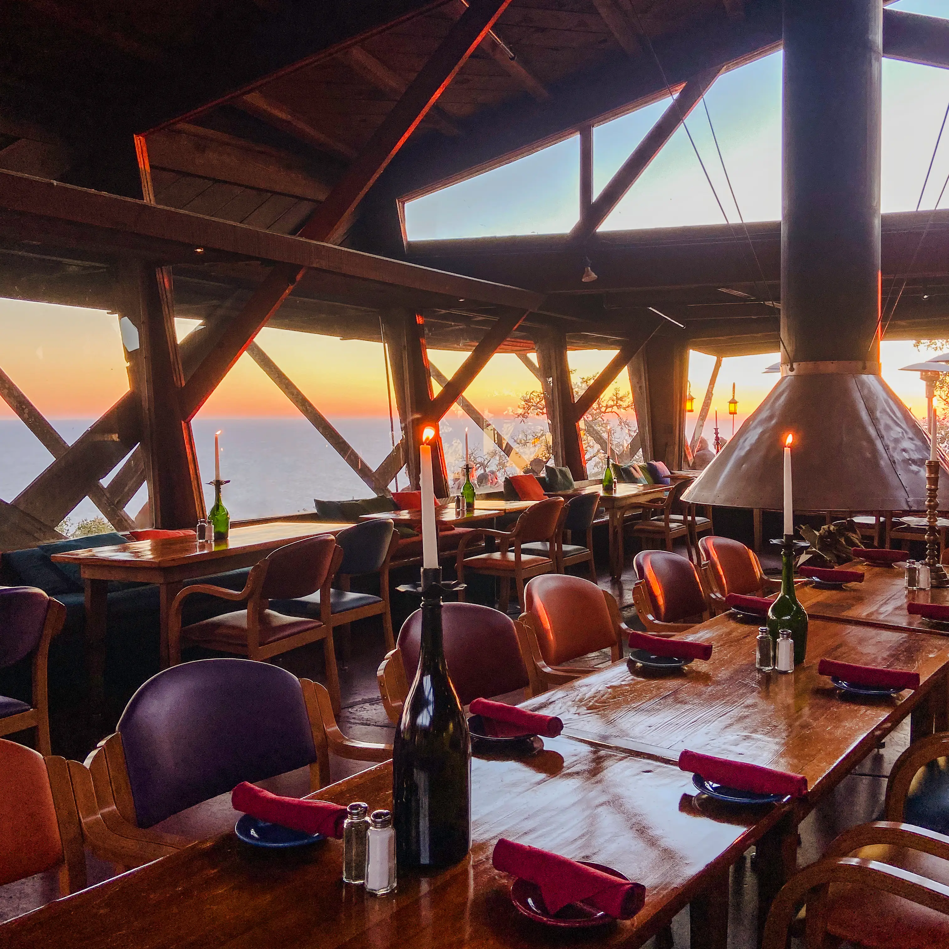 Wooden restaurant dining room with set tables, candles, and large windows overlooking the ocean at sunset.