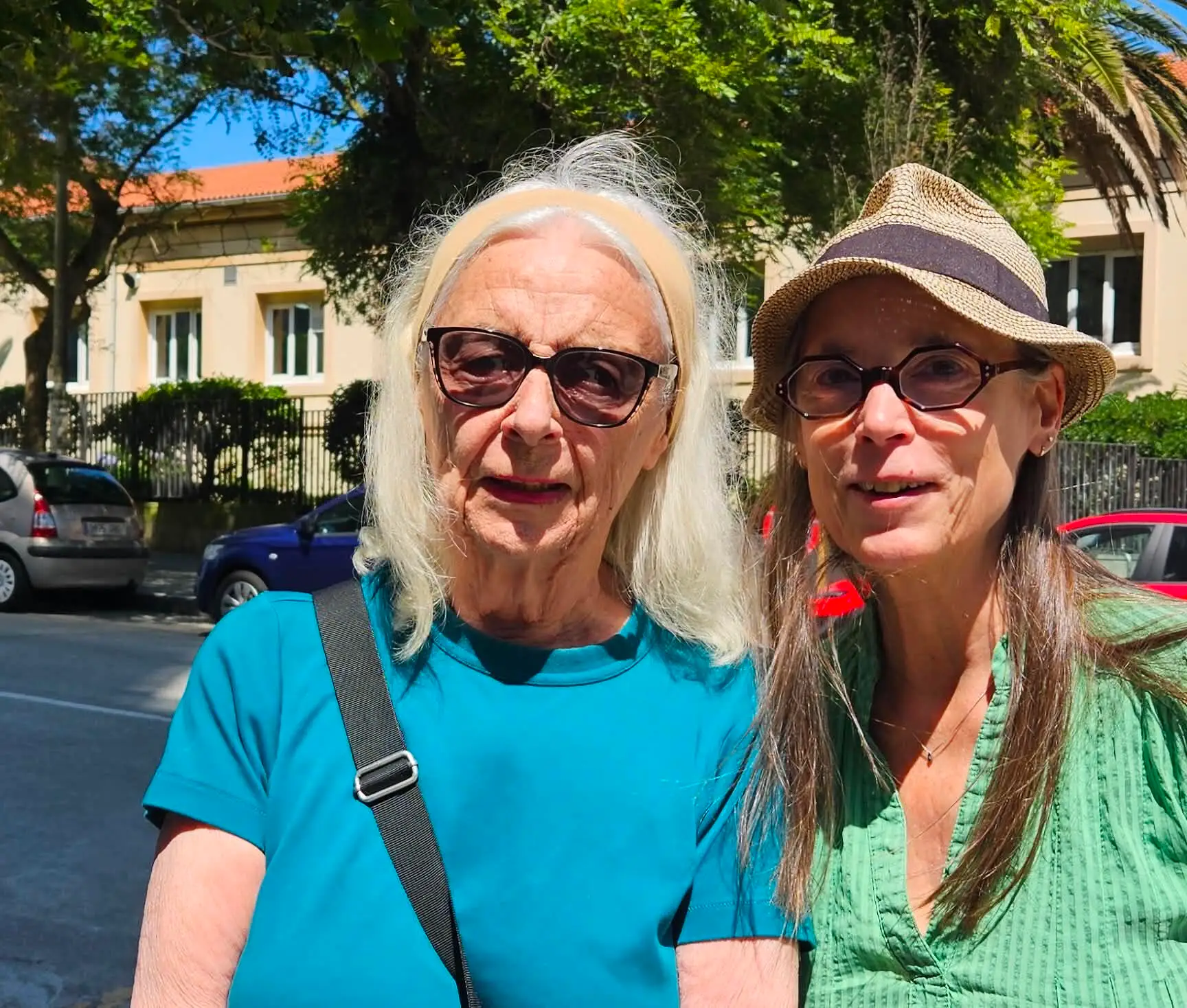 Two people wearing sunglasses stand together on a sunny street with parked cars and trees behind them.