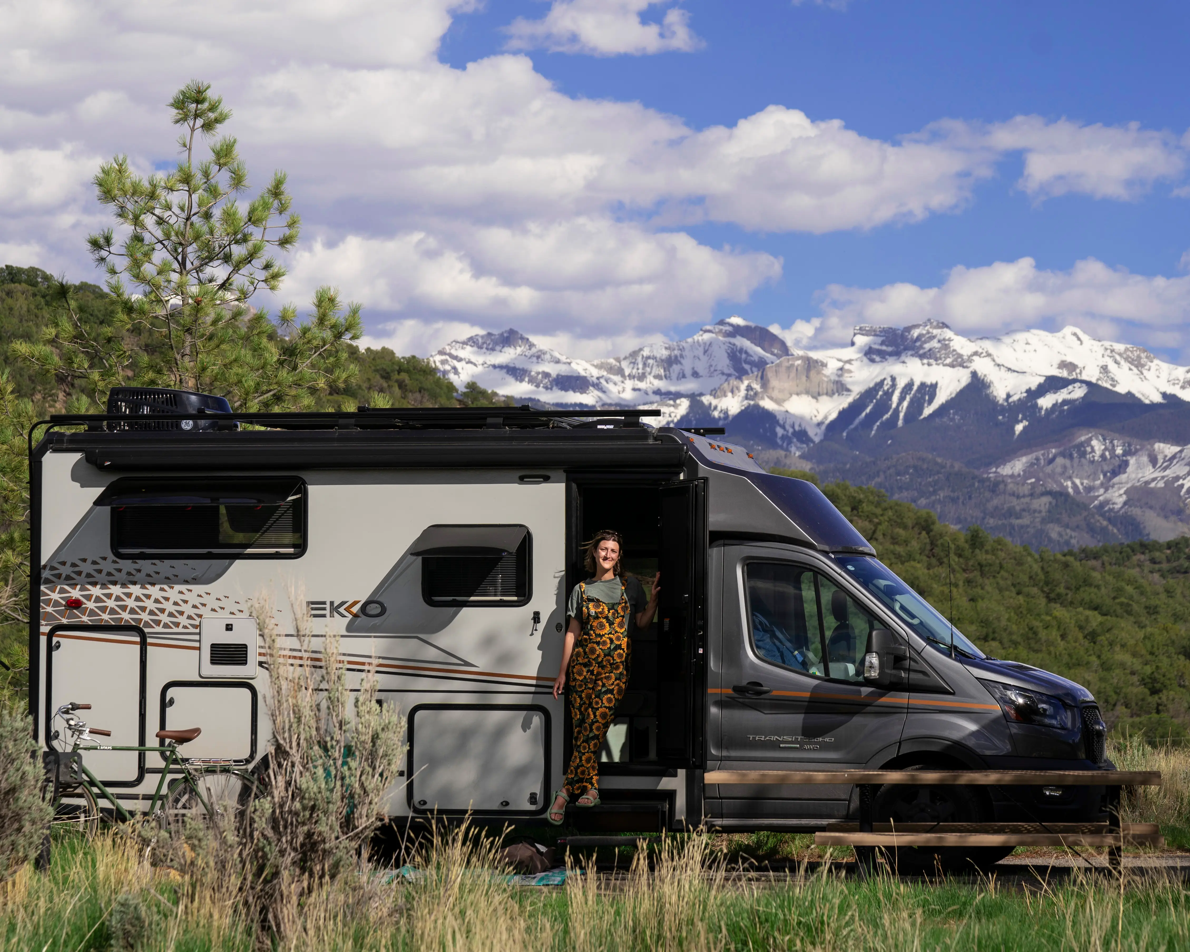 Sarah Silbiger standing in a camper van doorway beside a bicycle with snowy mountains and forest in the background.