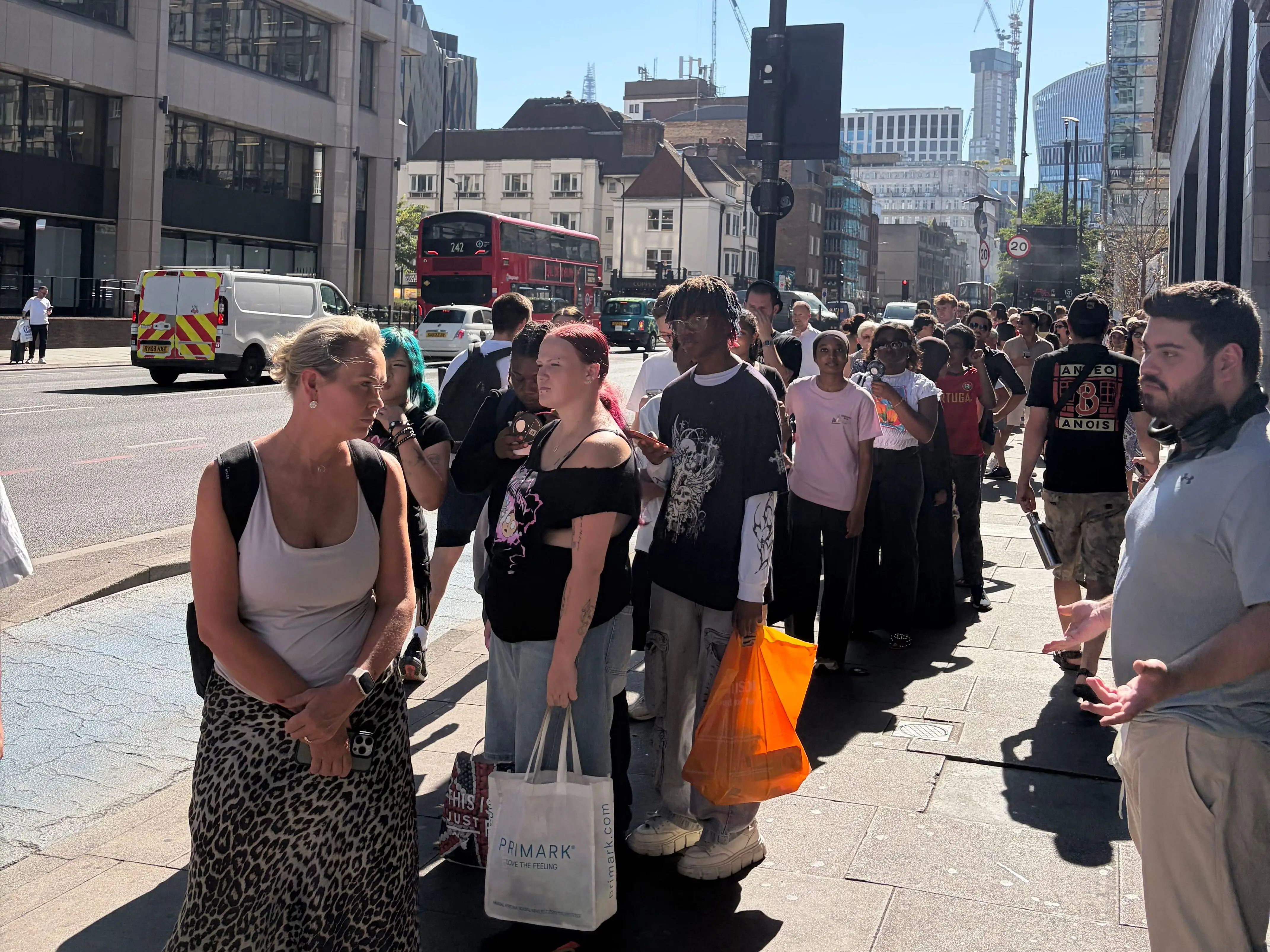 A long queue of people waits on a sunny city sidewalk beside traffic and tall office buildings in London.