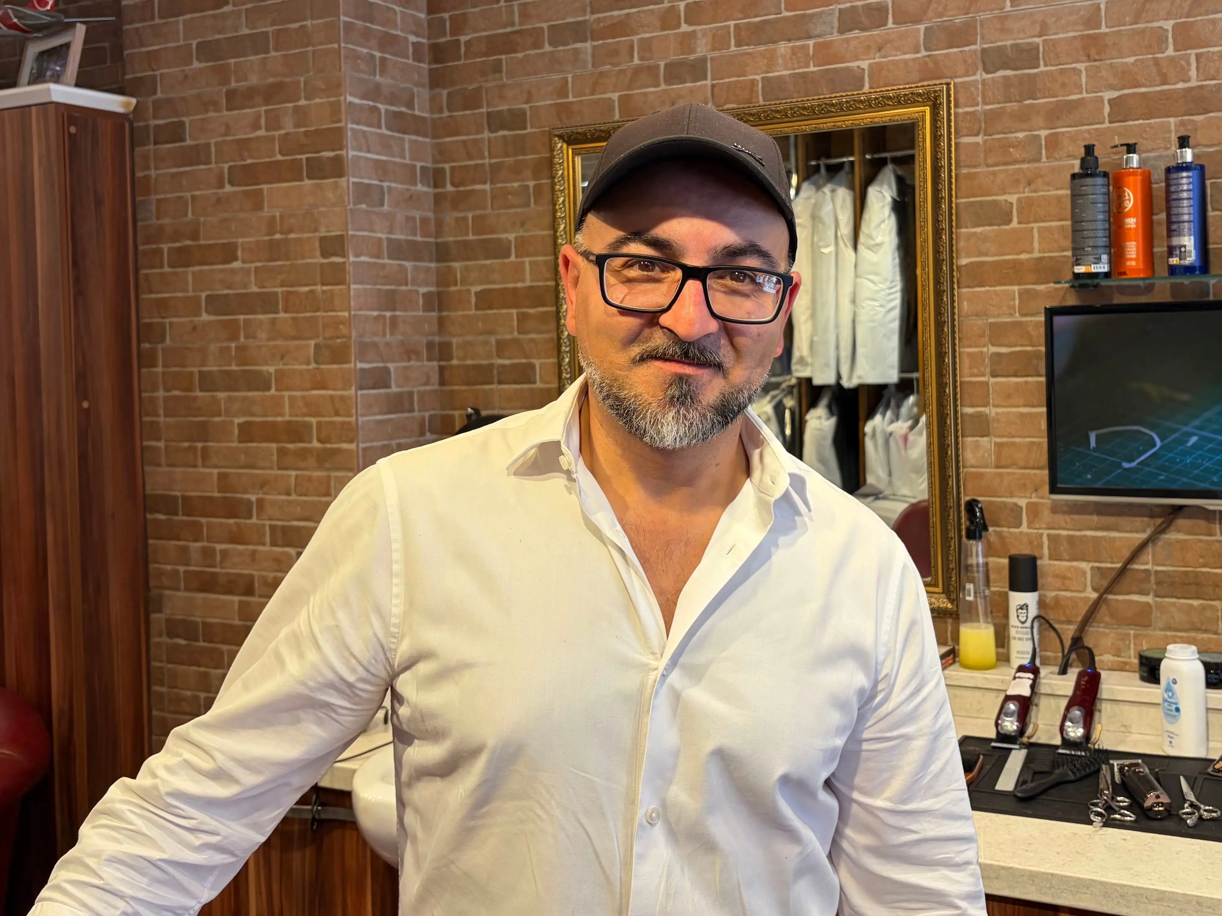 Person in glasses stands inside a barbershop with brick walls, mirrors, grooming products, and barber tools.