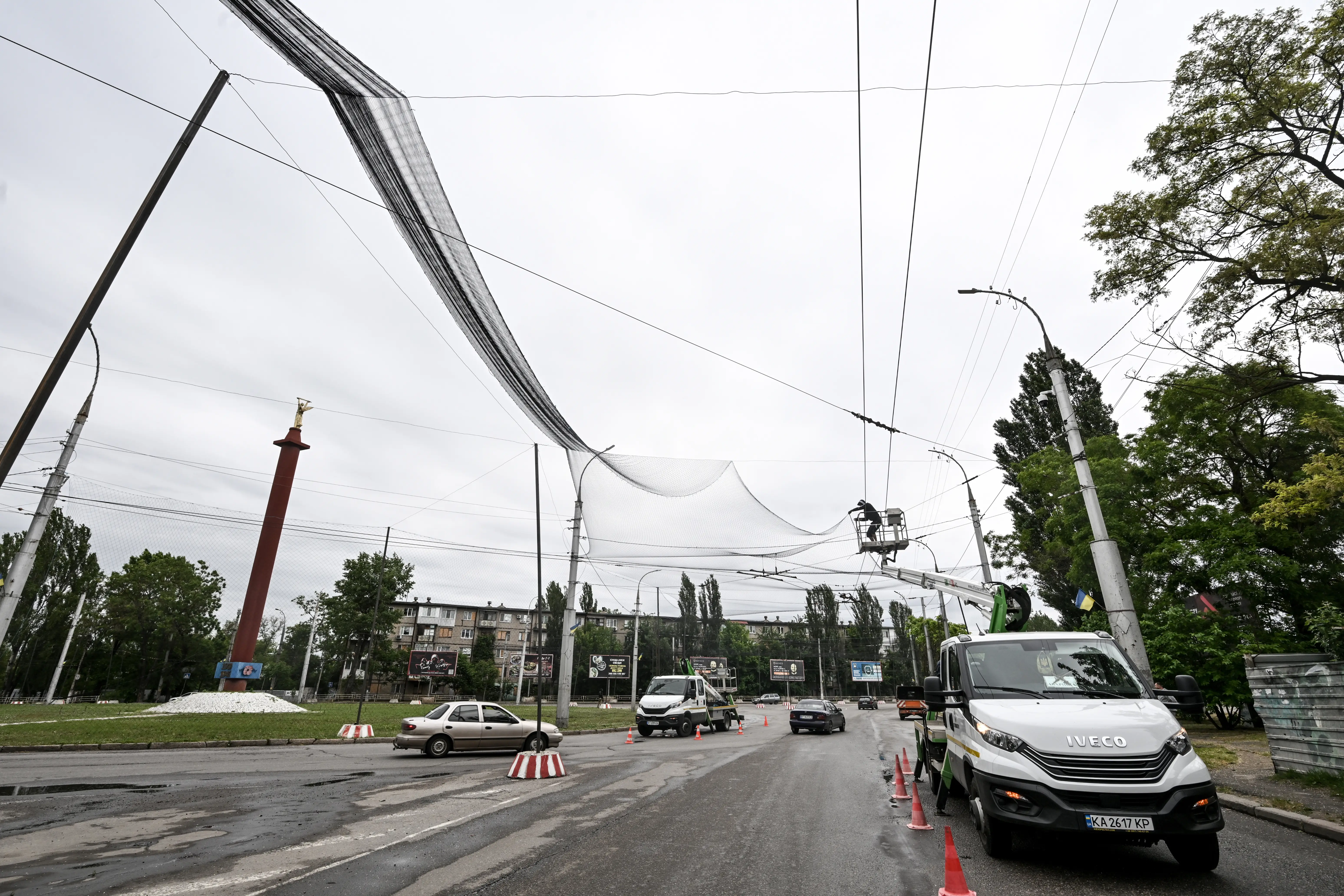 Utility workers on a bucket truck install anti-drone nets above an overcast urban road with traffic cones.
