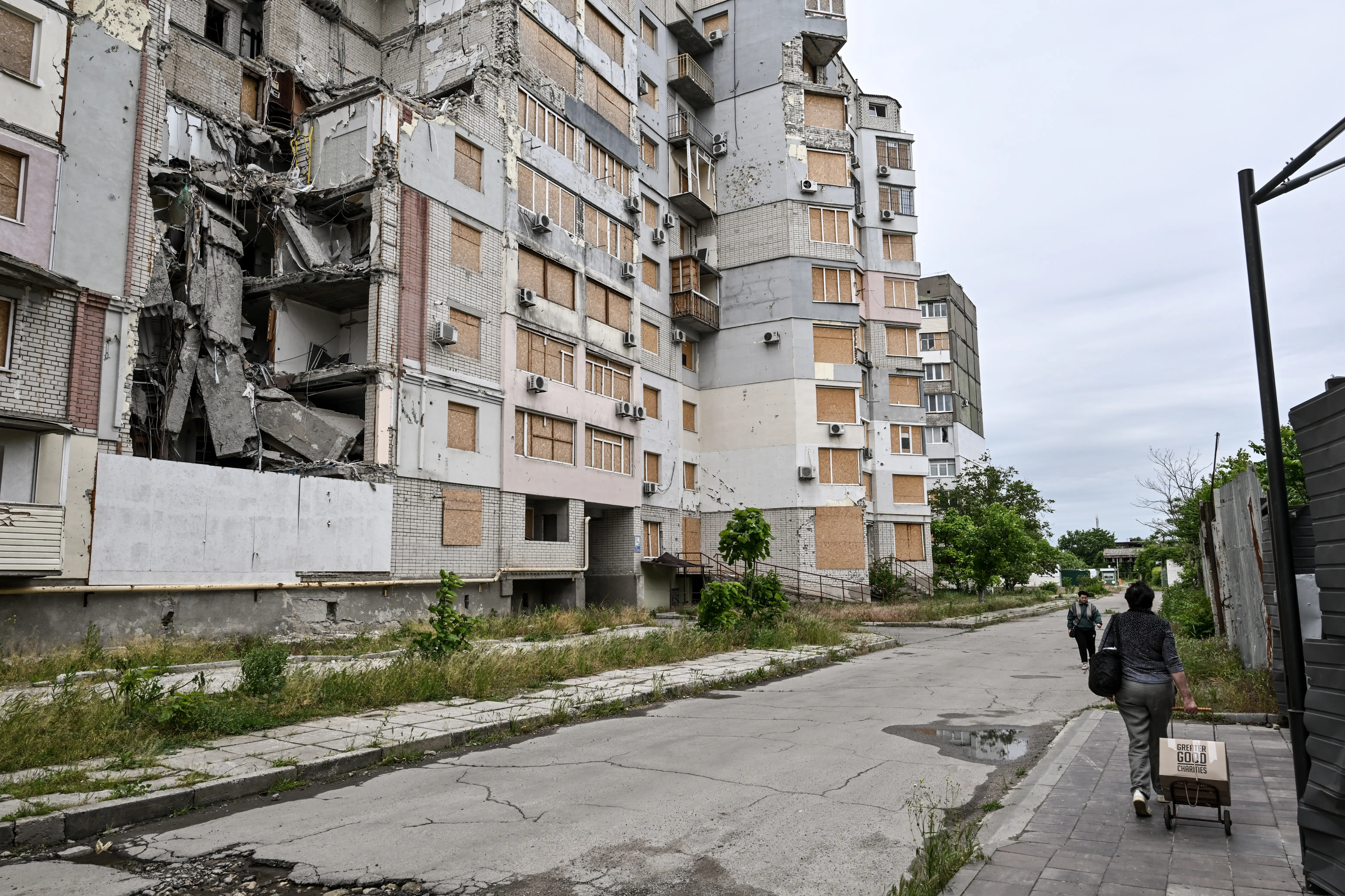 Damaged apartment block with collapsed sections and boarded windows stands beside a cracked street with pedestrians.