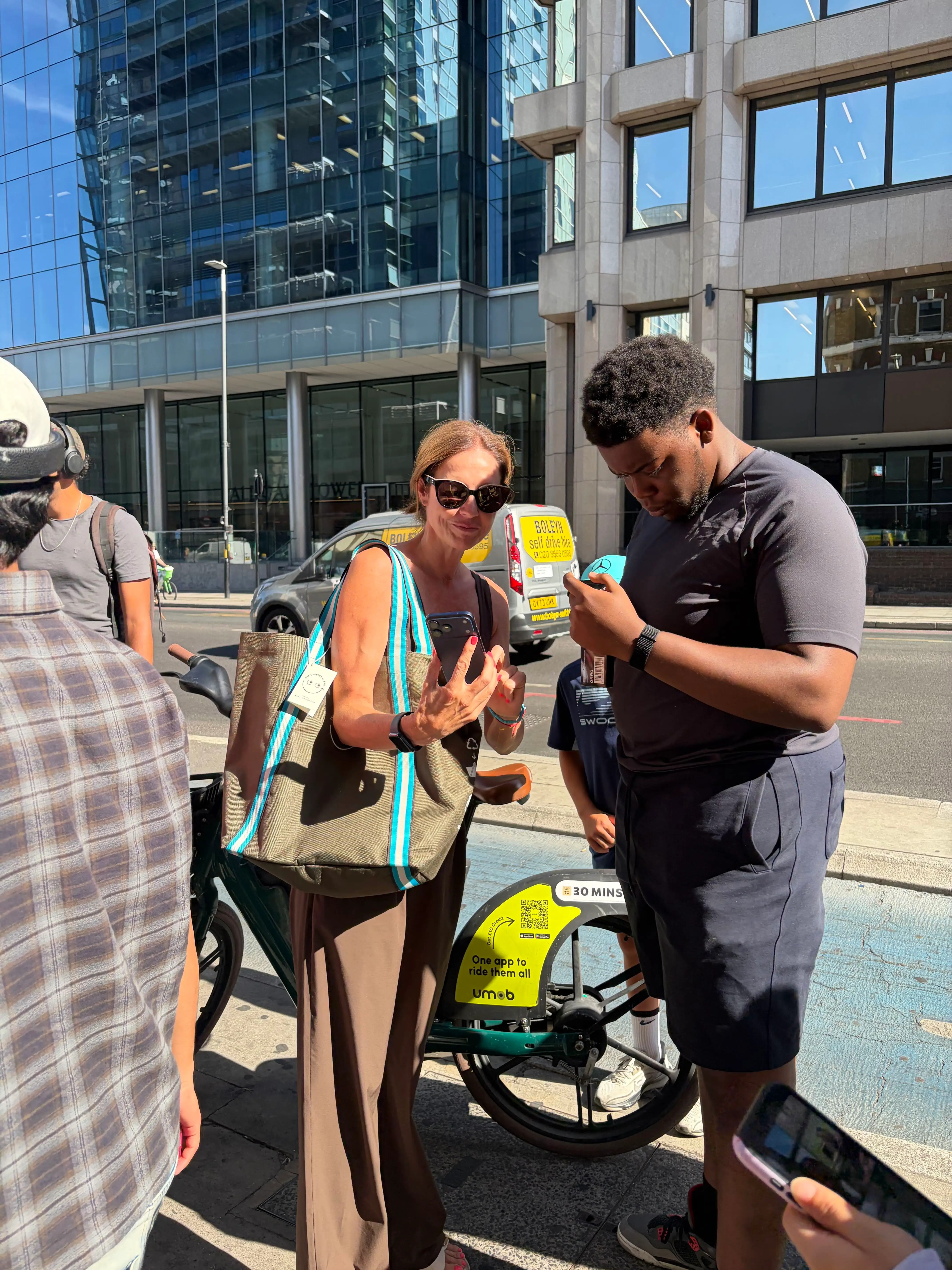 People use smartphones beside a rental bike on a sunny city street outside modern office buildings.