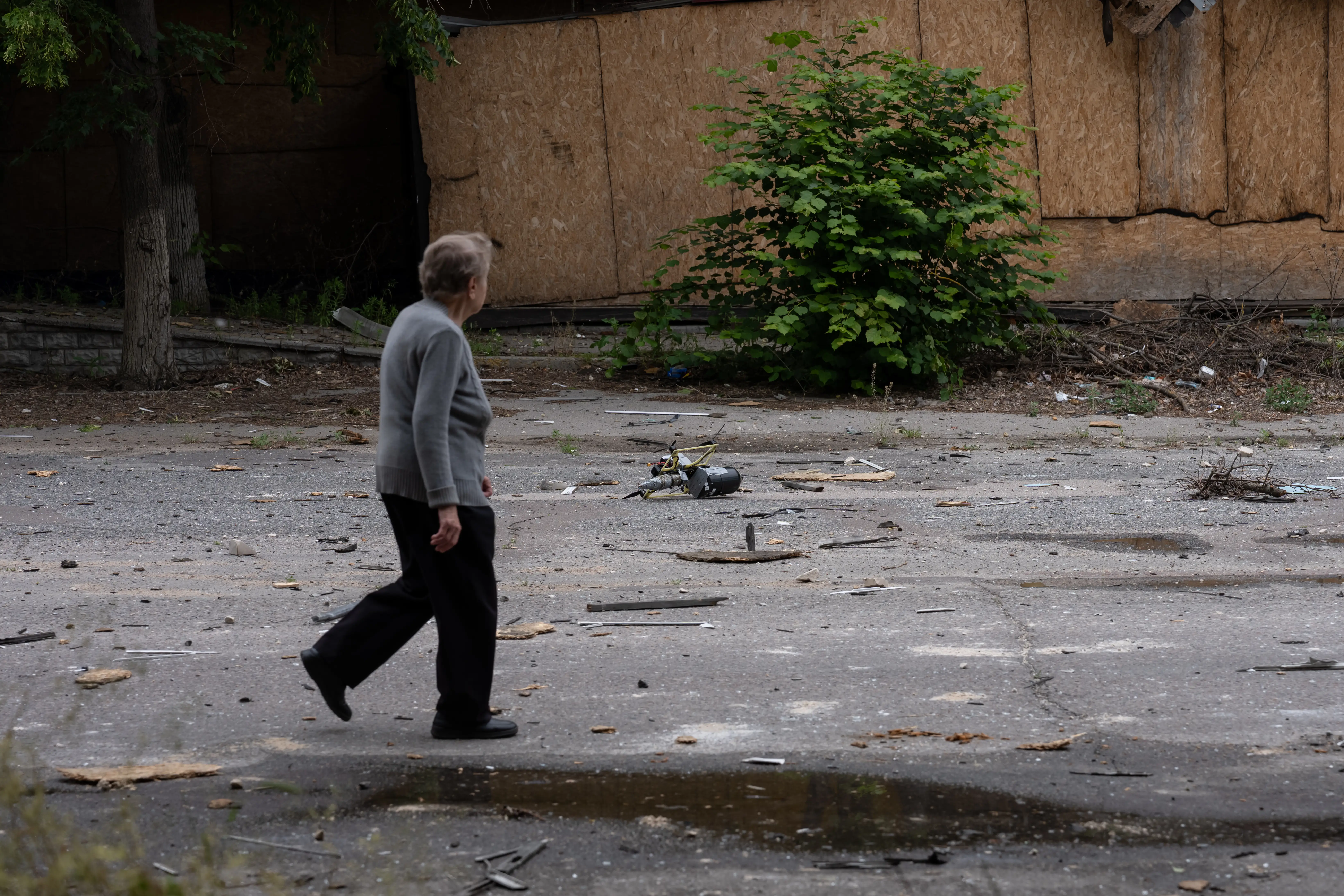A person walks across a debris-strewn paved area beside a boarded-up building and overgrown shrubs. A drone lies on the ground.