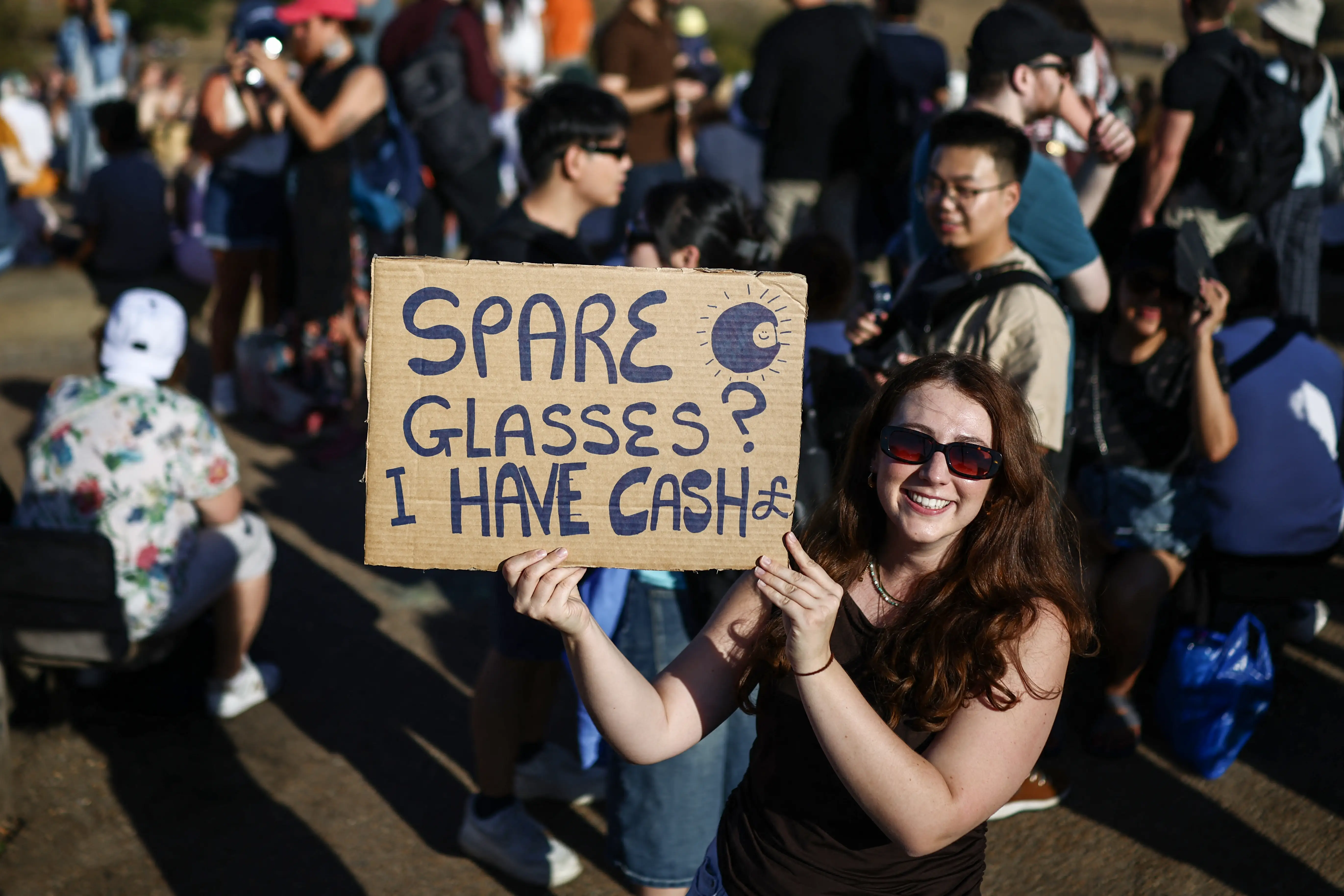 A woman holds up a cardboard sign asking for solar eclipse sunglasses.