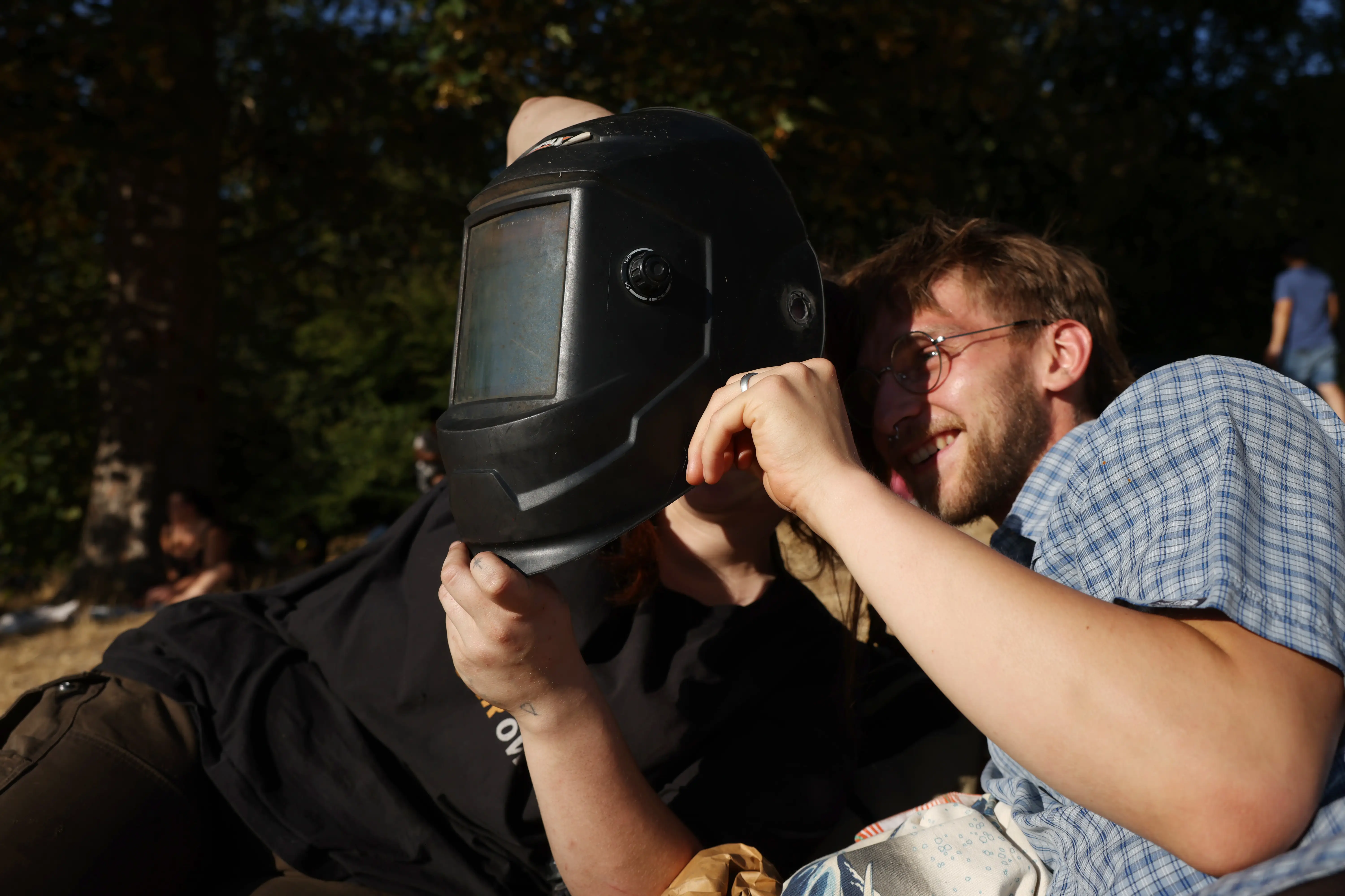 Two people sitting down hold up a welder's mask to view the solar eclipse.