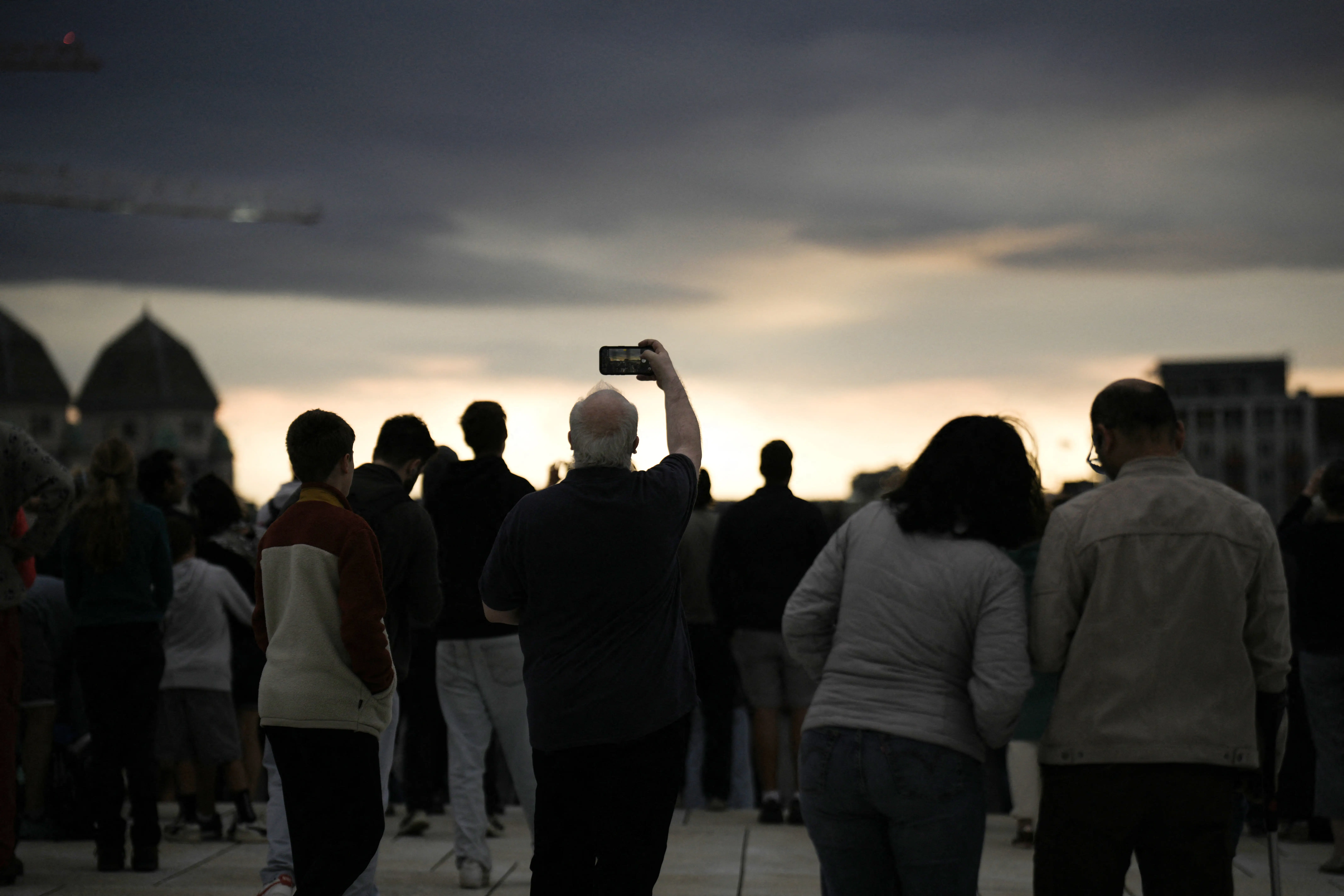 A group of people stands on a rooftop, looking up at a gray sky.