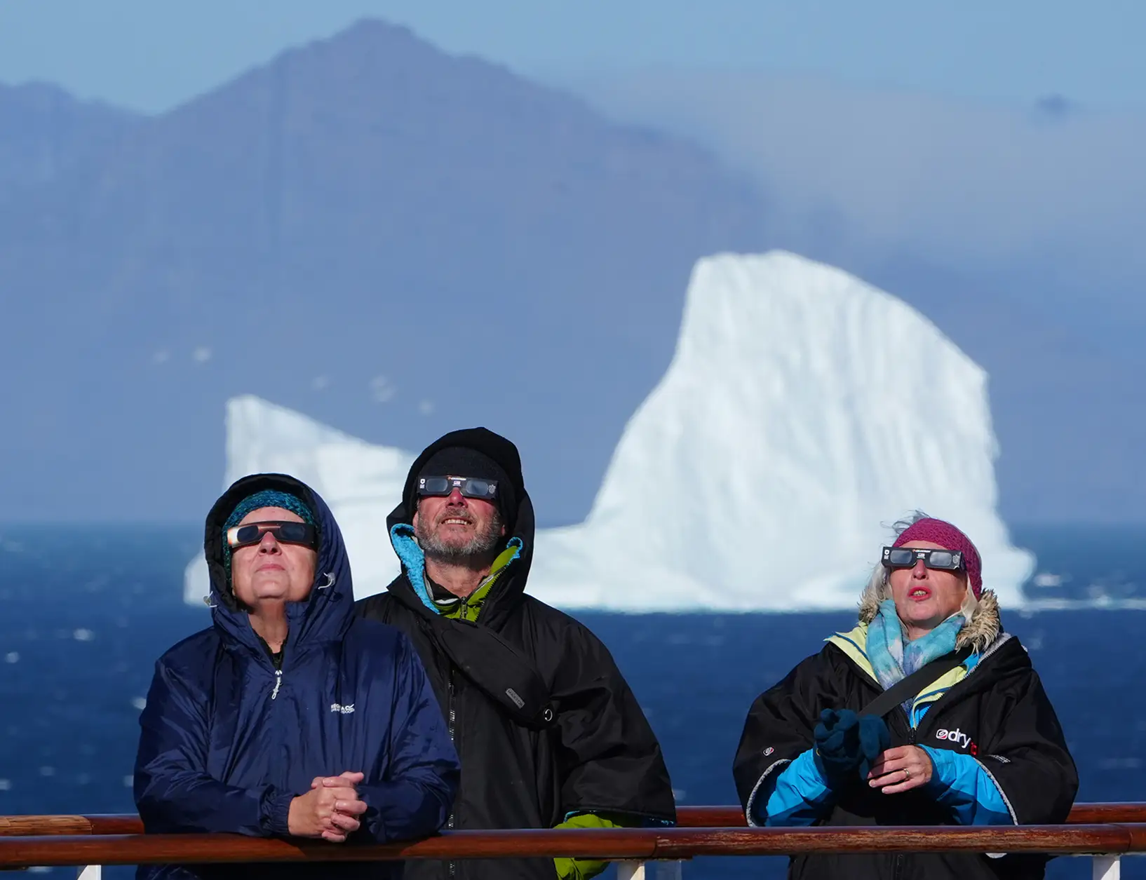 Three people in windbreakers look up at the sky, with a large iceberg in the background.