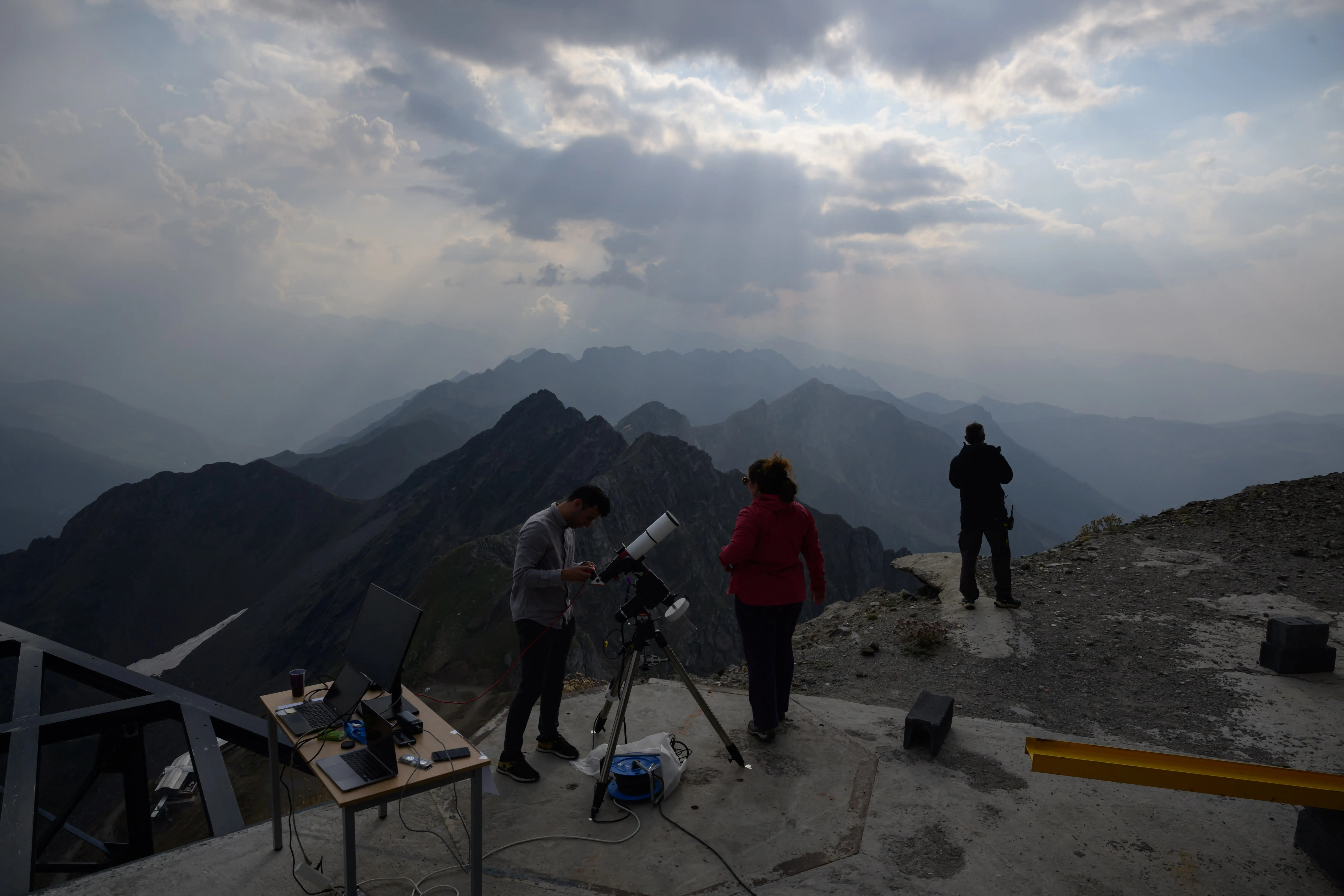Three people stand on top of a mountain with science equipment situated on a concrete slab.