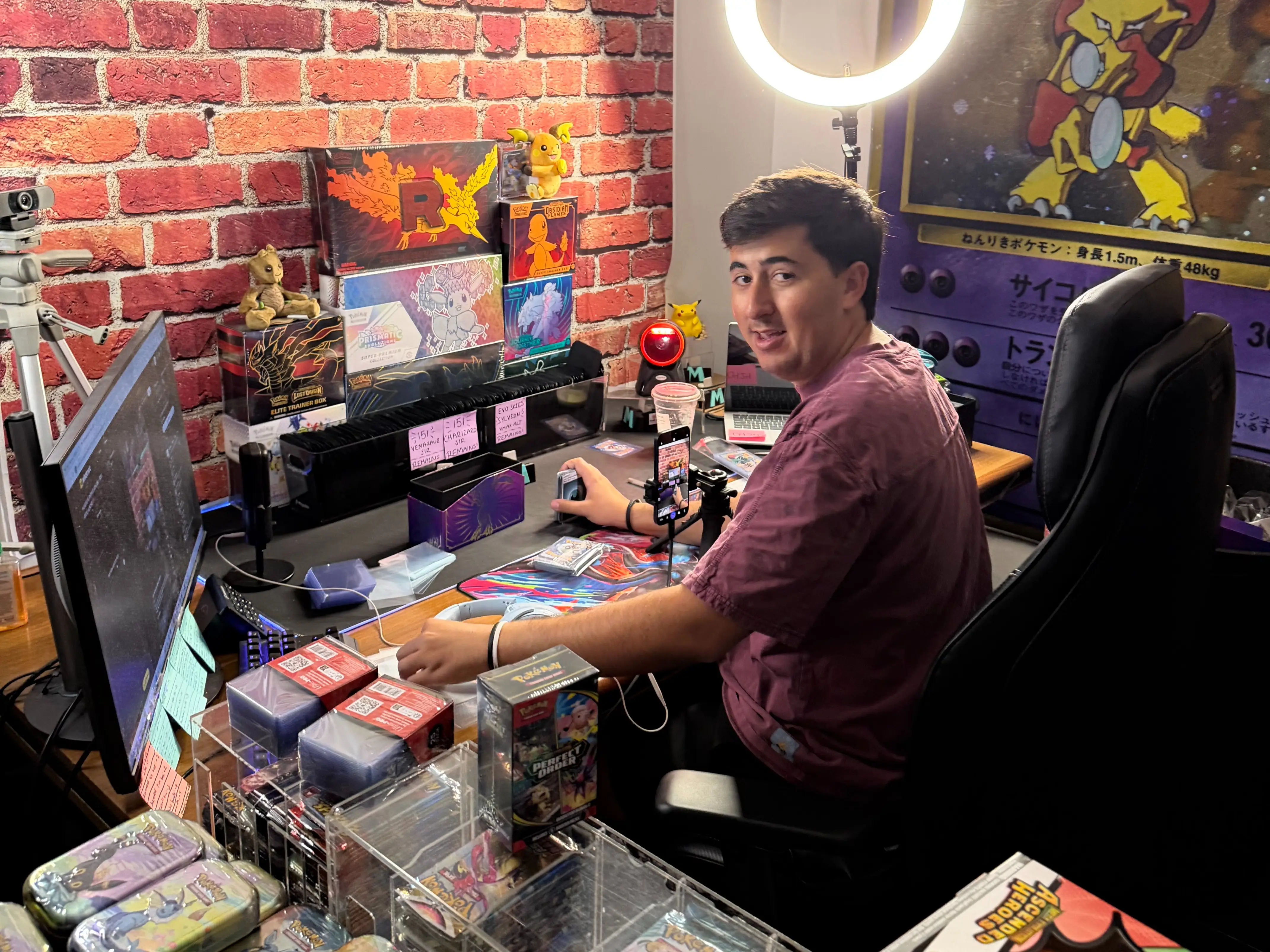 Person seated at a Pokémon-themed trading card workspace with camera, ring light, displays, and card supplies.