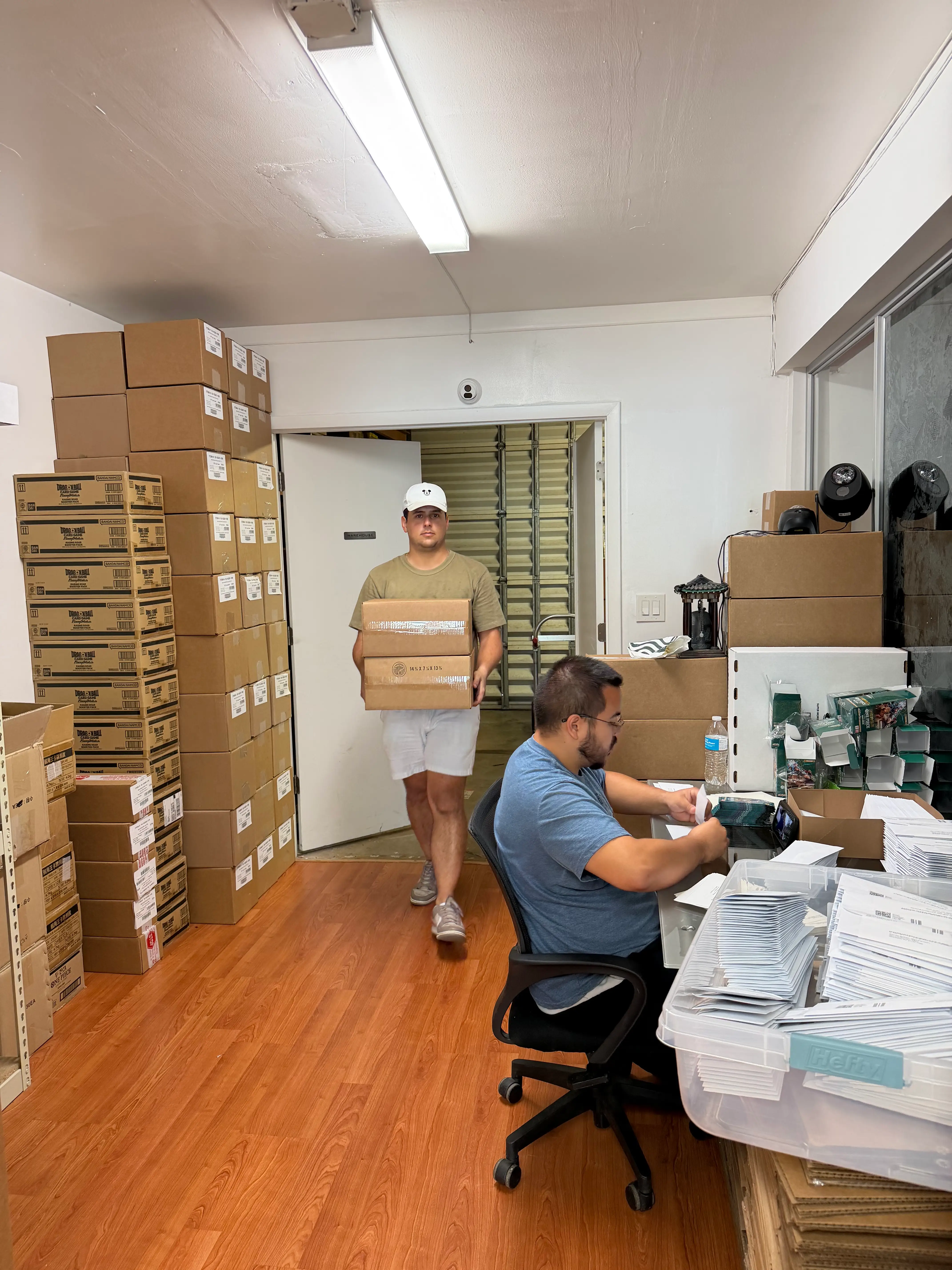 Two people work among stacked cardboard boxes and mailers in a small shipping and packing room.