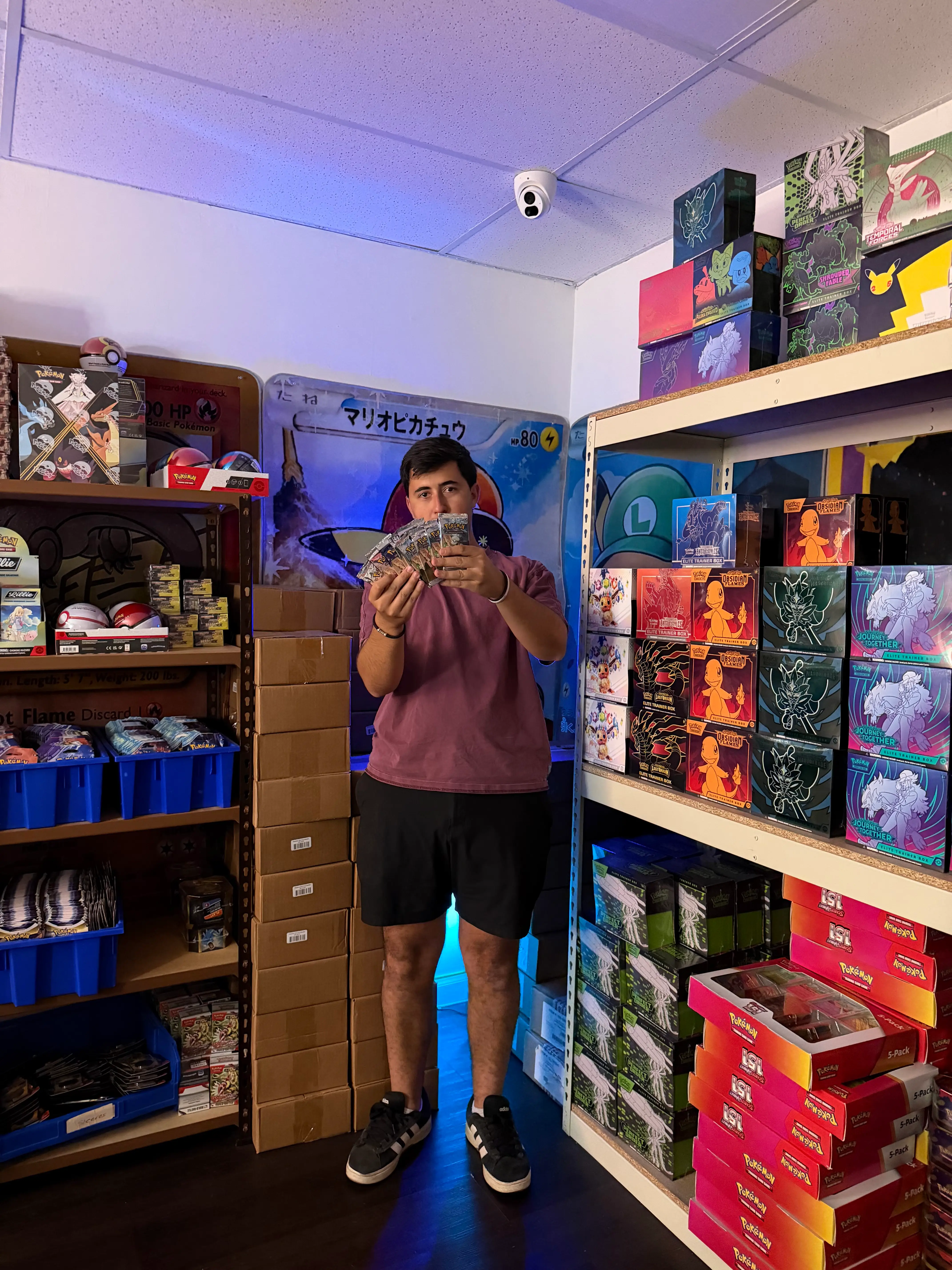 Person holding trading cards stands between shelves of Pokémon merchandise in a collectibles shop.