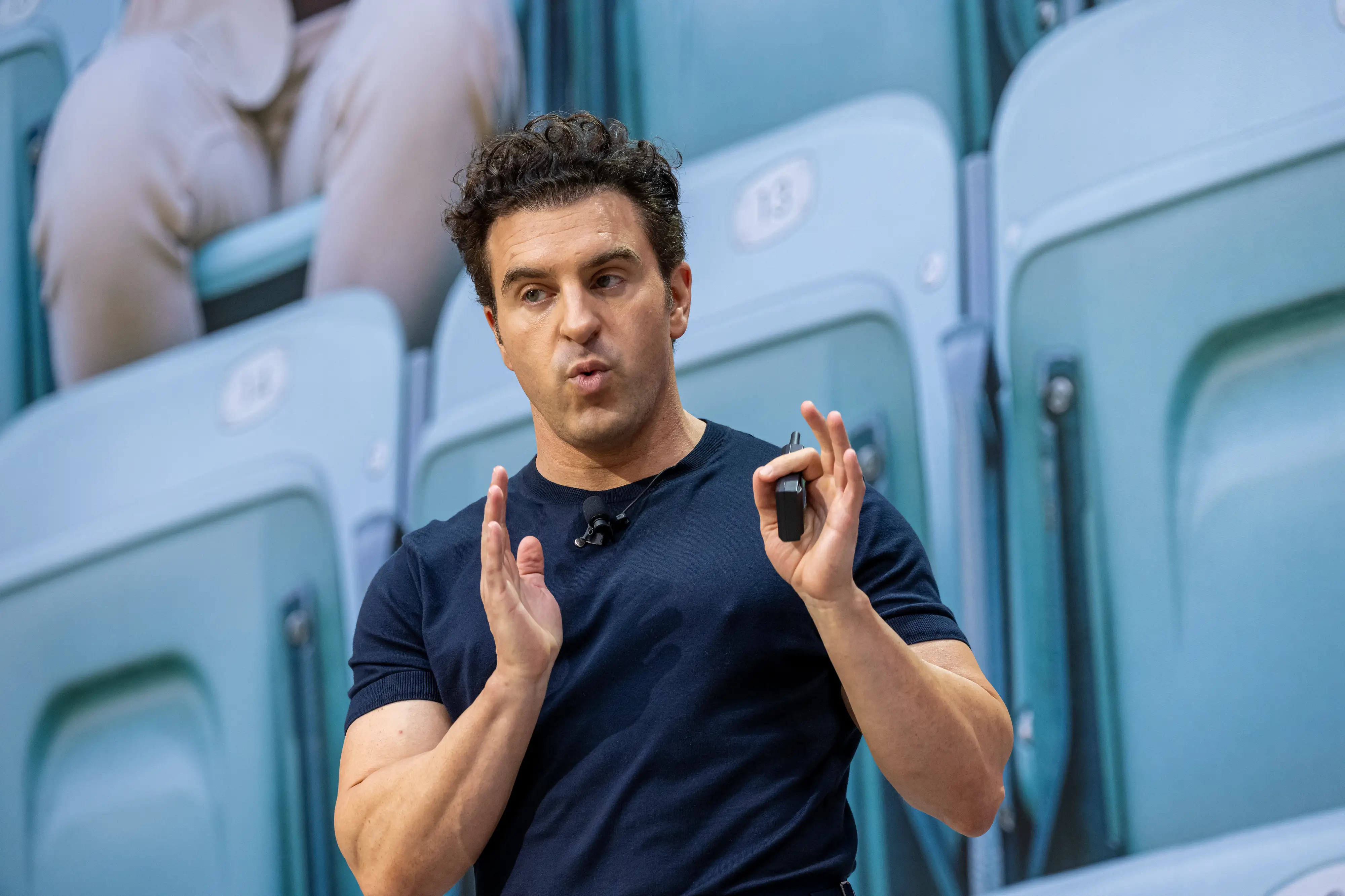 Brian Chesky in a dark shirt gestures while holding a remote in front of blue stadium seats.