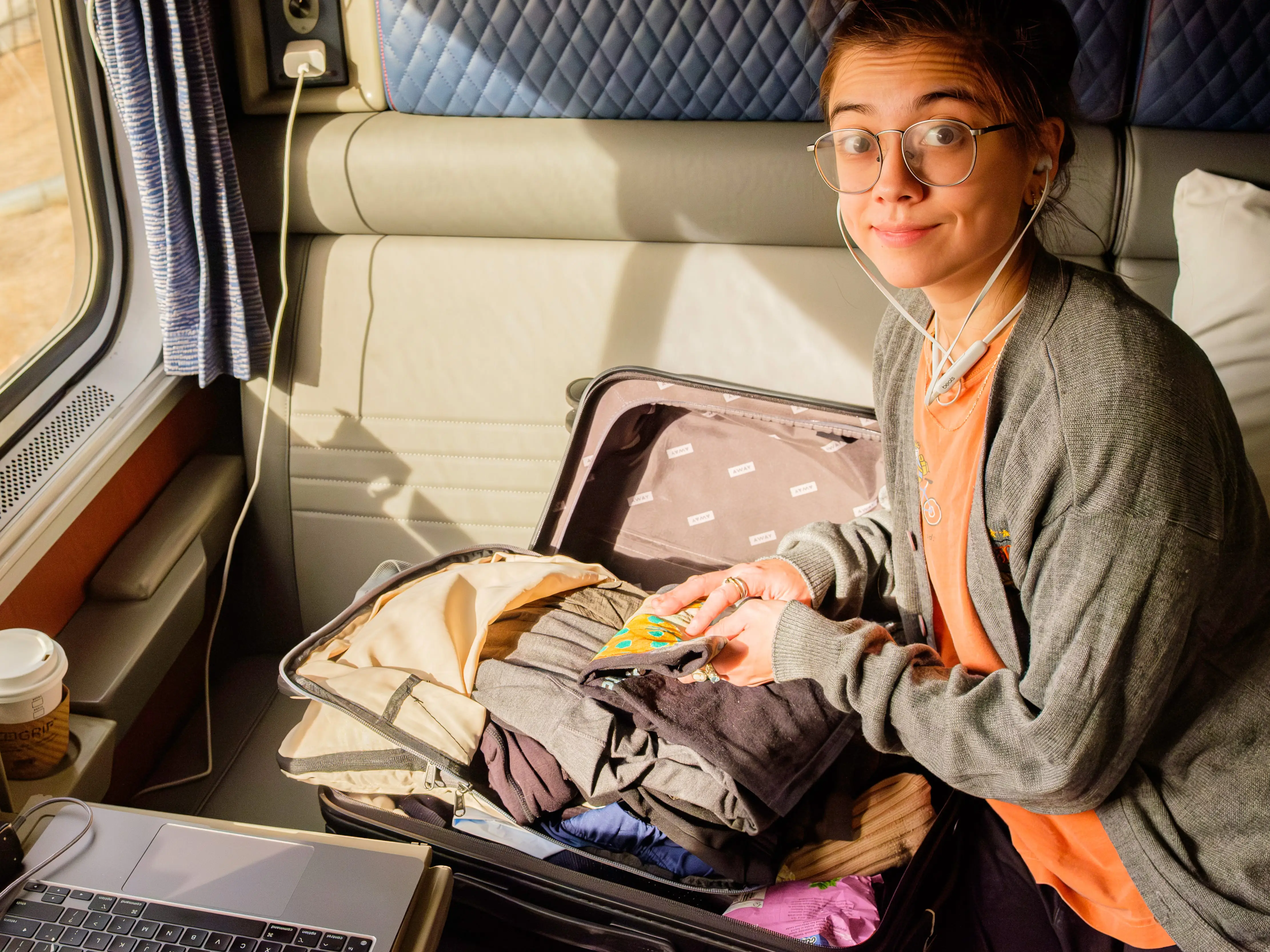 Traveler wearing earbuds sits in a train compartment beside an open suitcase, laptop, camera, and window.