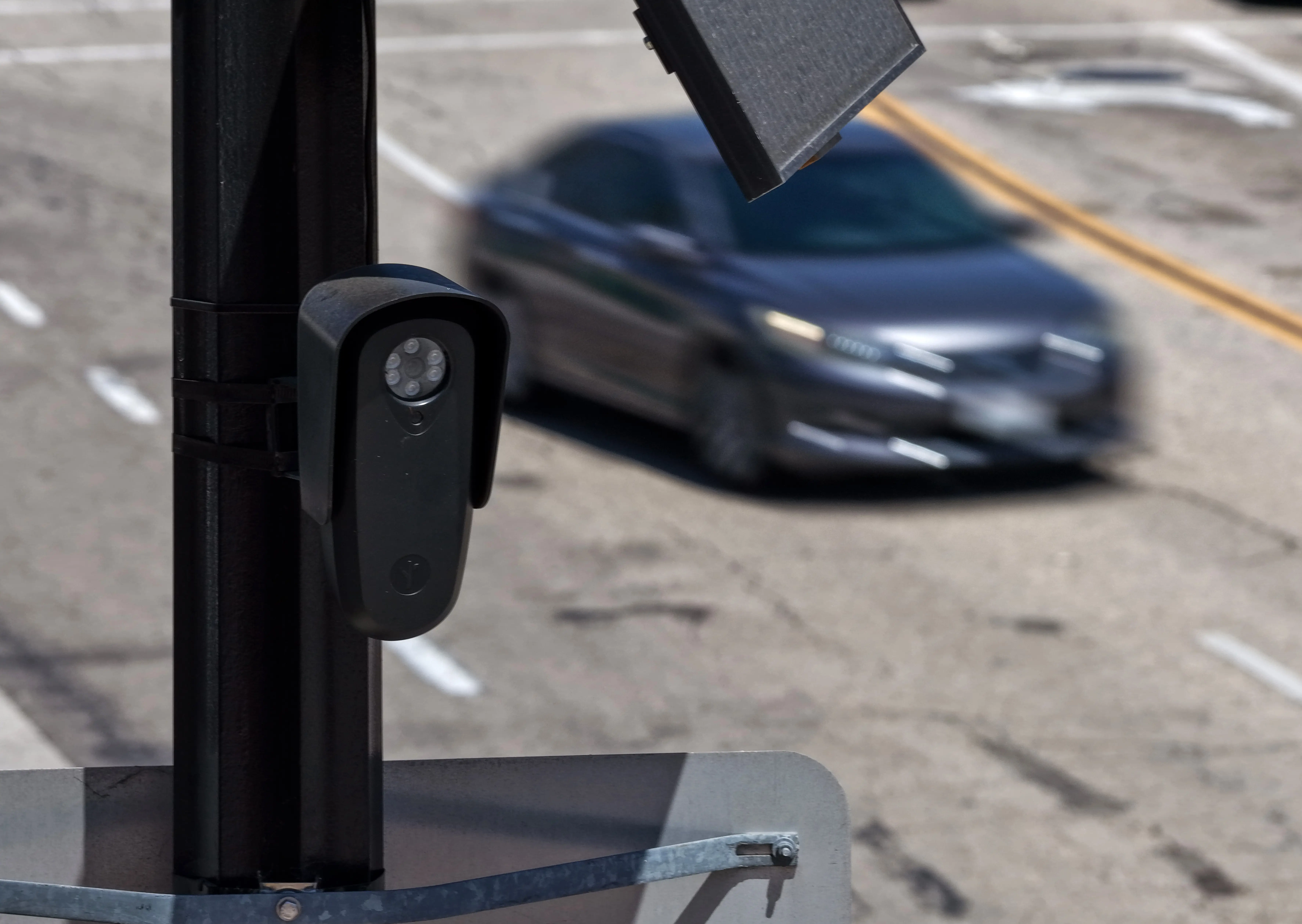A Flock camera monitors a roadway while a gray sedan passes by.