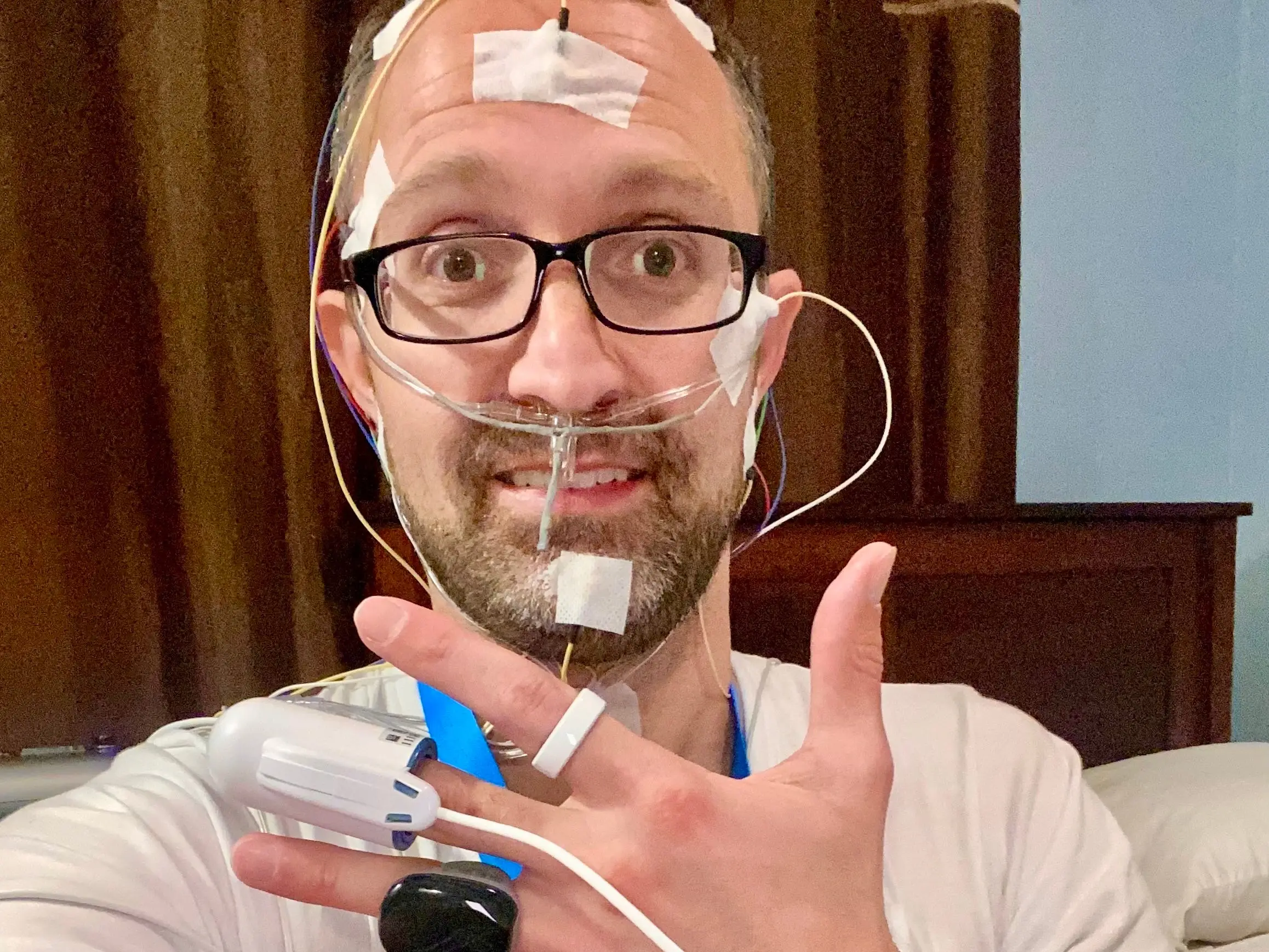 Dustin Freckleton testing a Happy Sleep device against traditional sleep-testing equipment, wearing glasses with tubes on his face and hand.