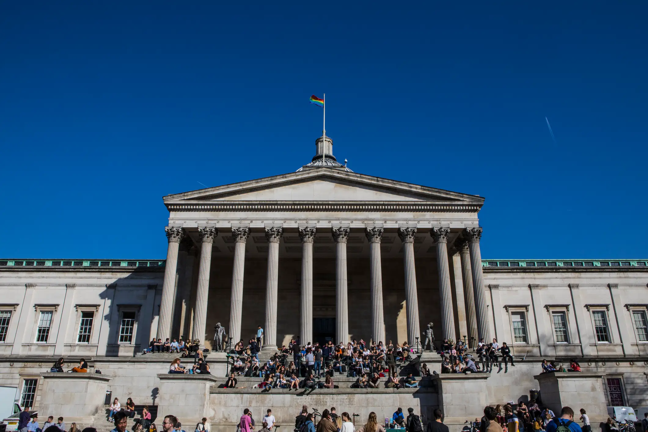The exterior of University College London with people sitting on the steps in front on a sunny day.