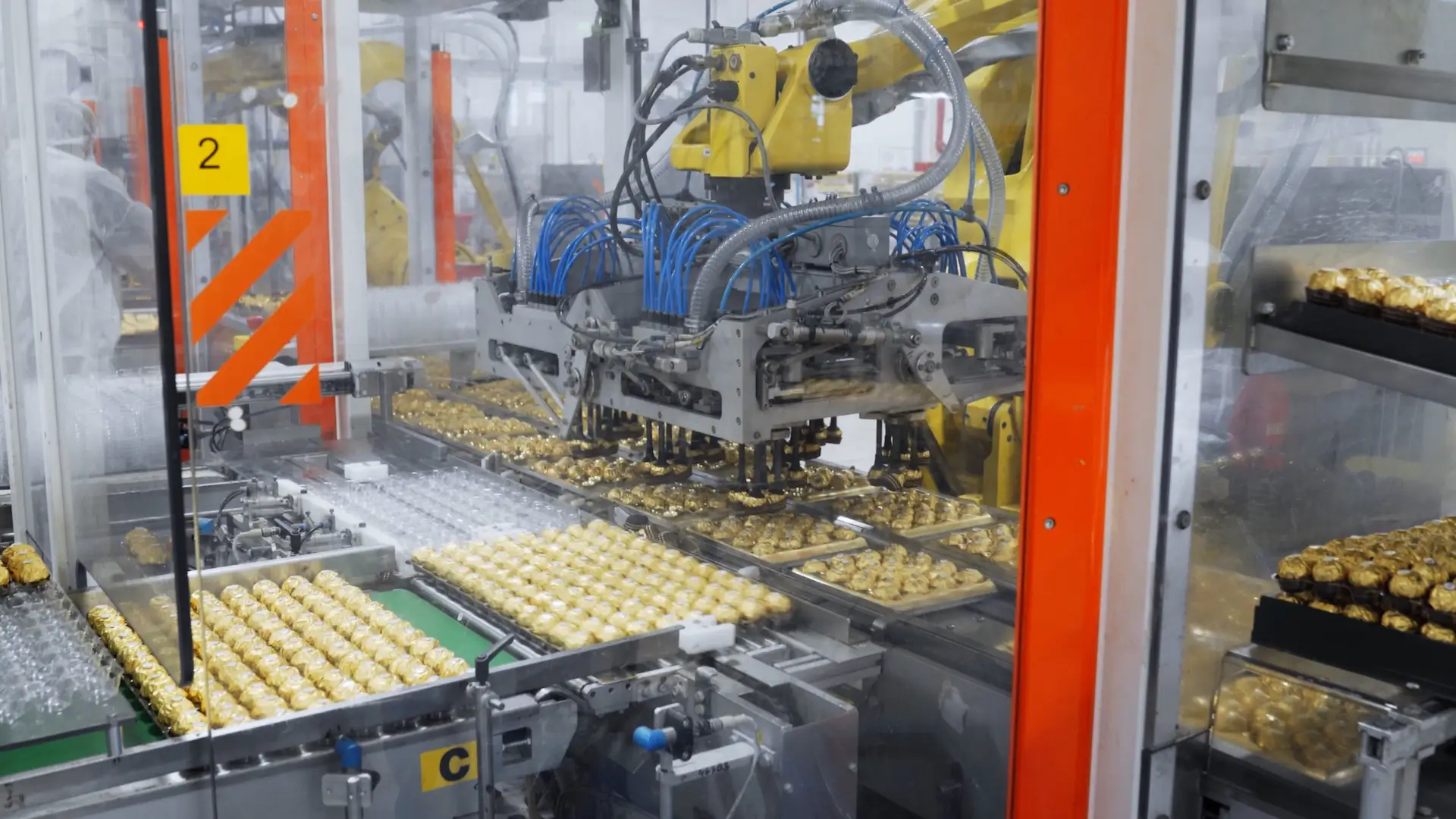 Factory shot of hundreds of golden Ferrero Rocher chocolates being sorted on a production line by large robots behind a protective glass screen.