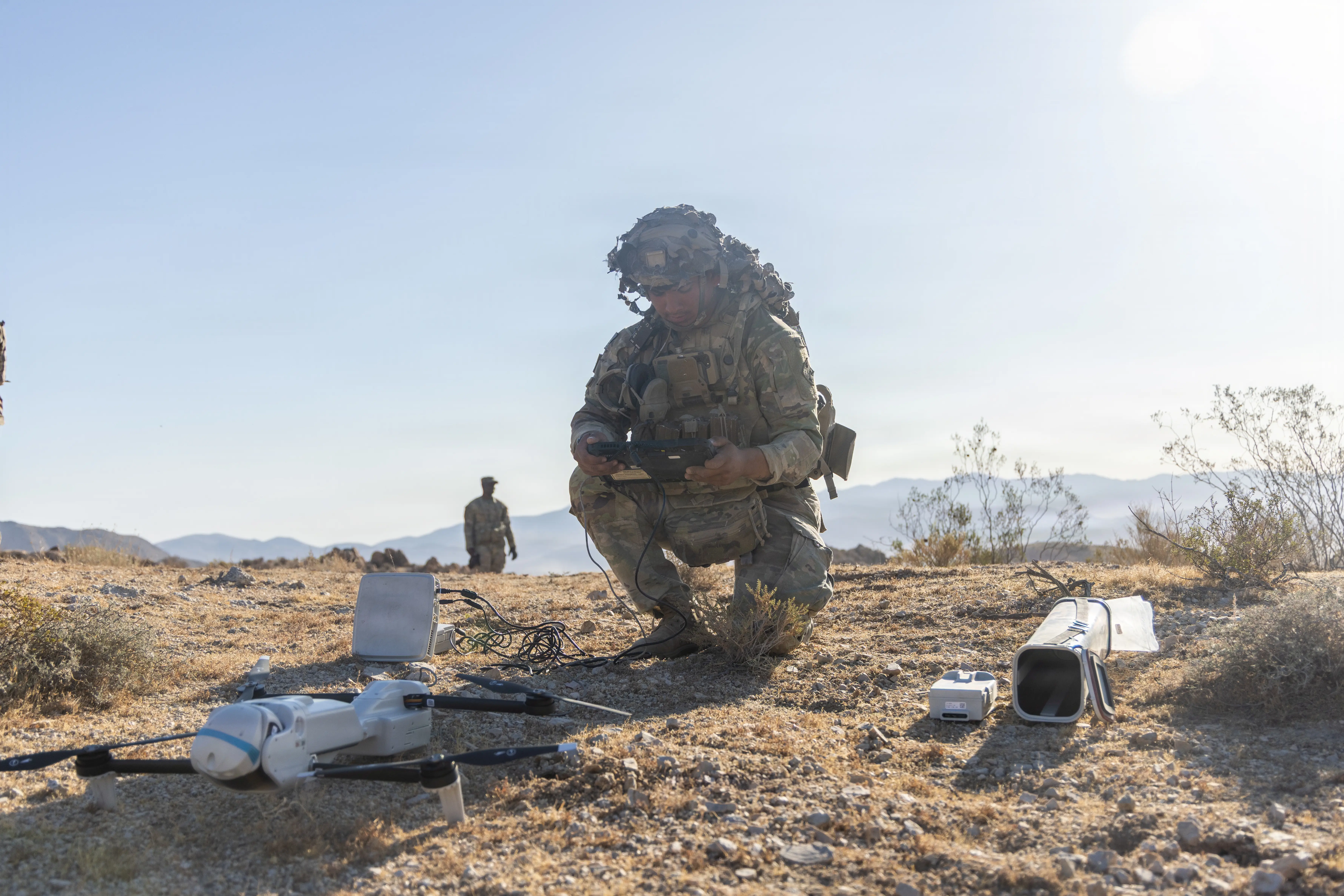 A US Army soldier crouches on the ground holding a drone controller. The drone and other drone technologies are placed in front of him.