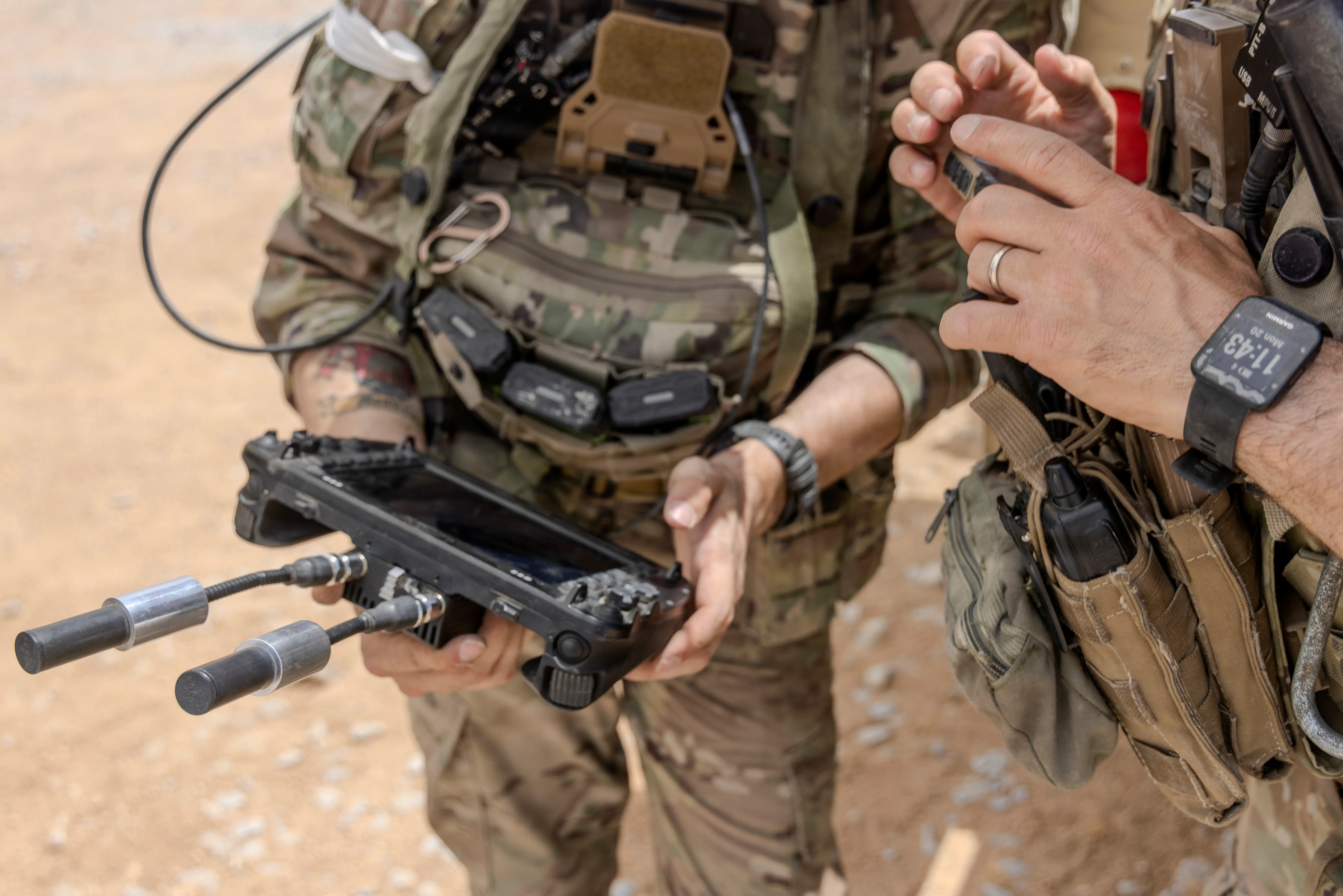 Two US Army soldiers stand in the desert. One is holding a drone controller.