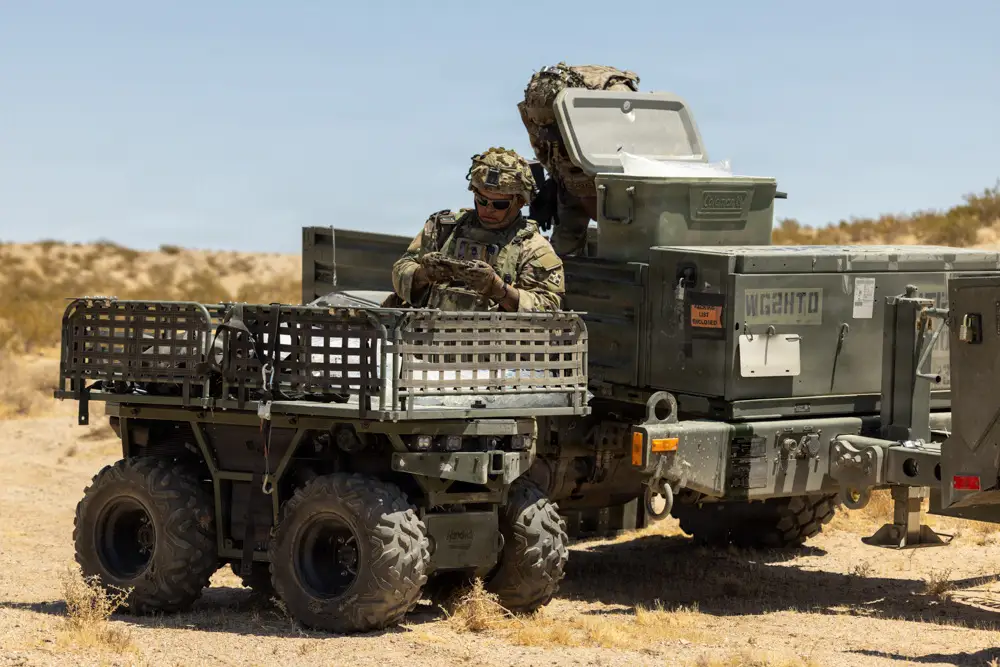 A US Army soldier stands alongside an uncrewed ground vehicle next to a truck in the desert.