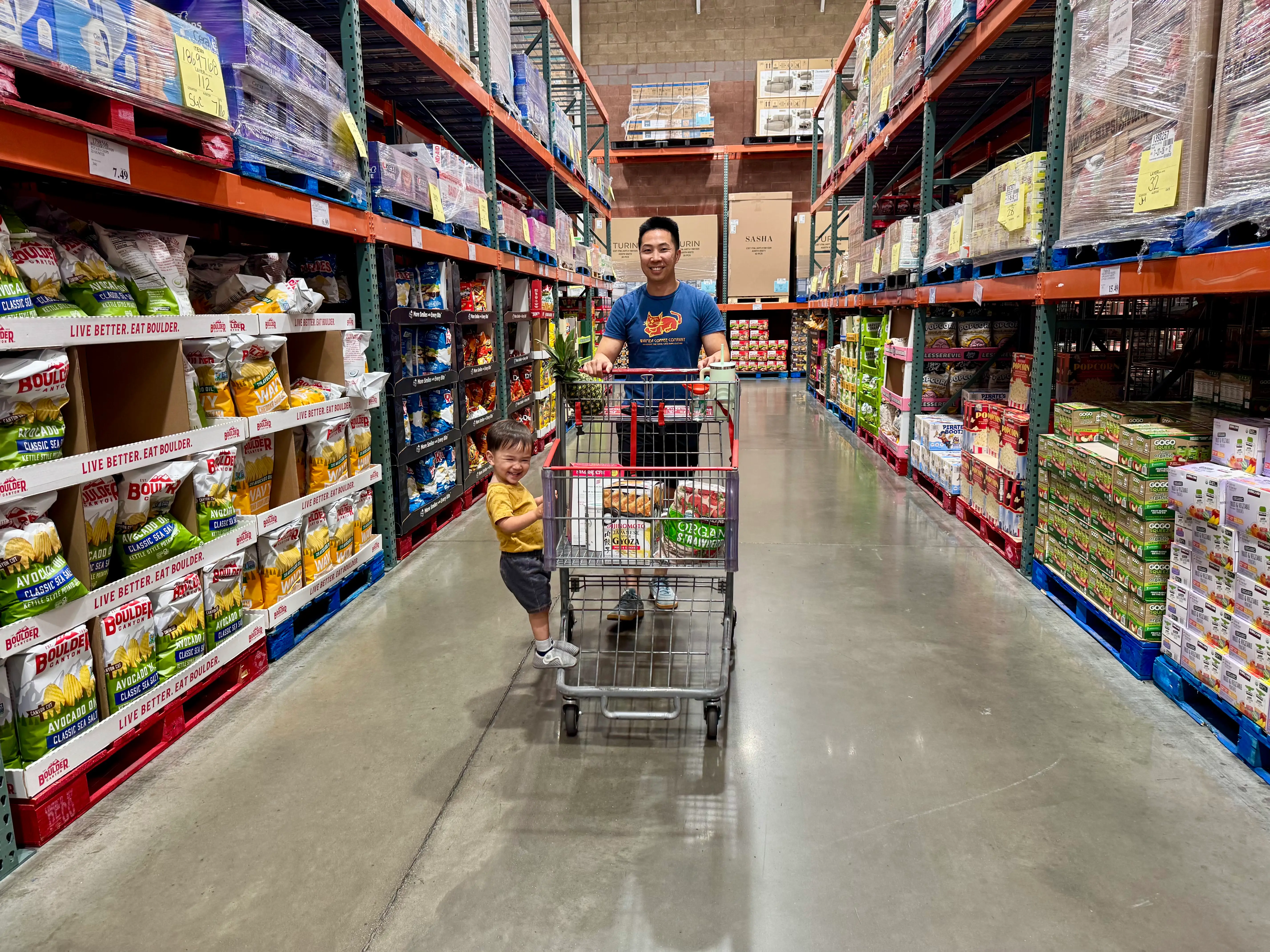 A man and a child rolling a filled stroller down a Costco snack aisle.