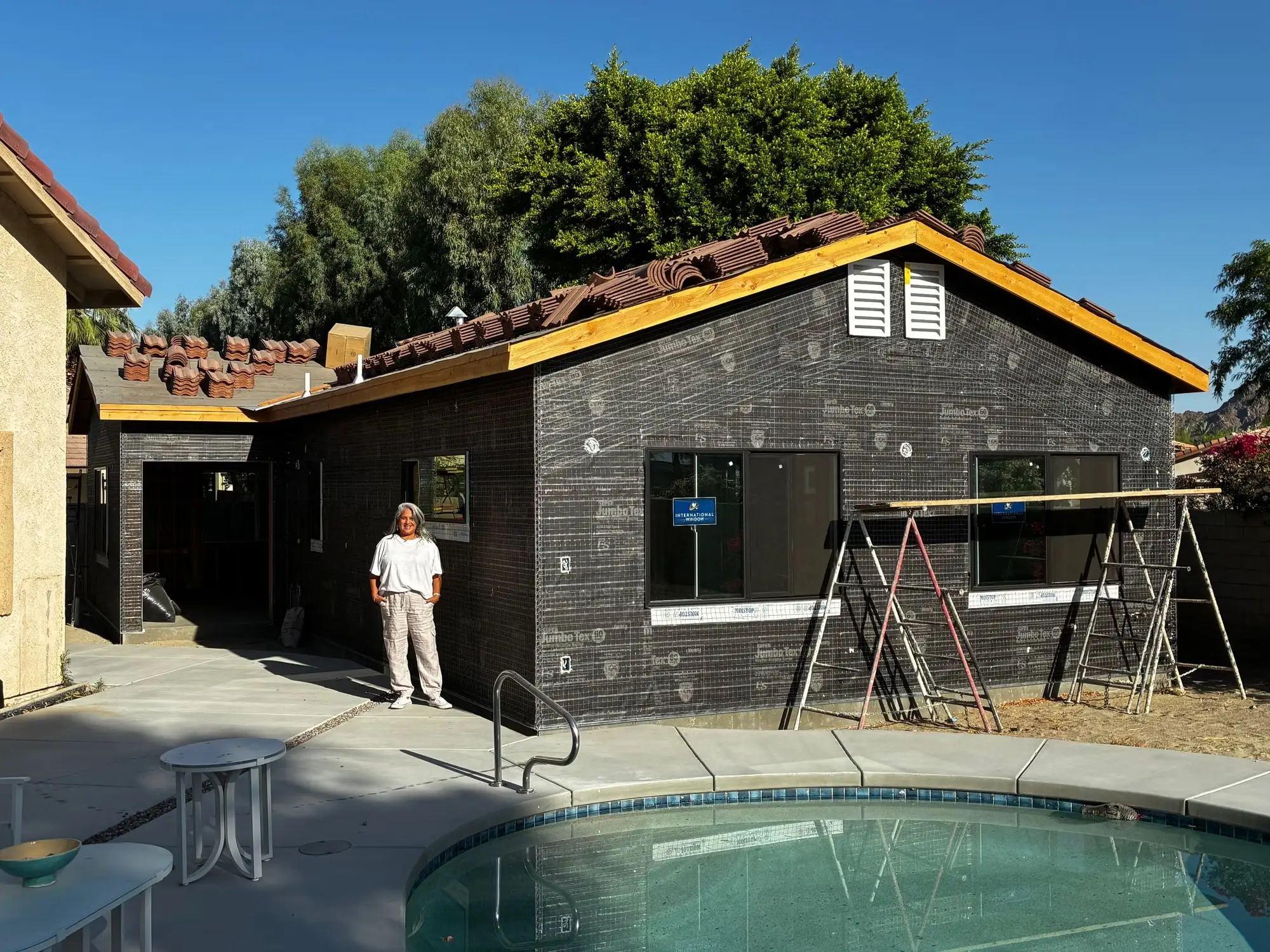 Person stands beside a backyard home renovation with exposed exterior wrap, scaffolding, roof tiles, and a swimming pool.