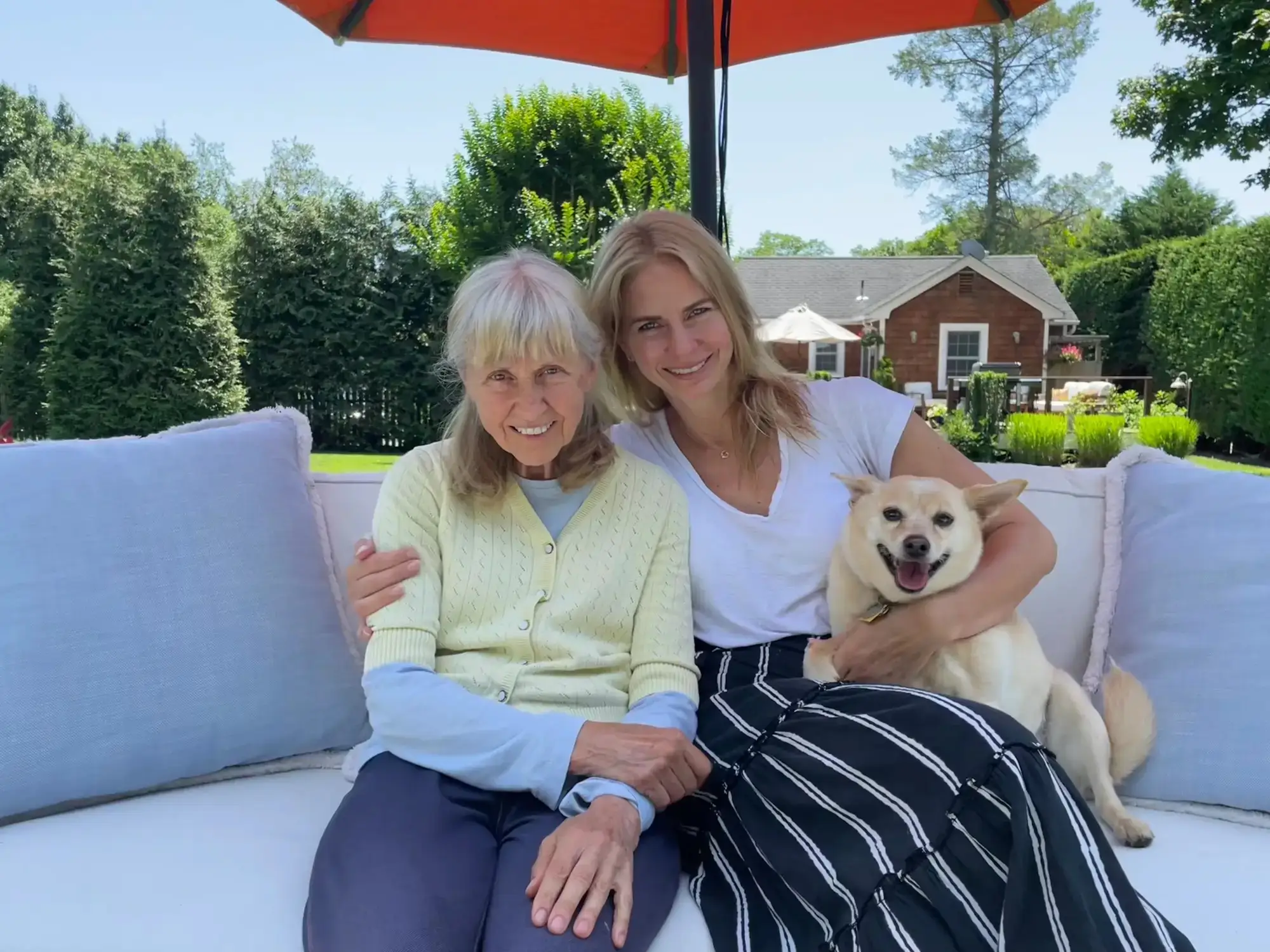 Lena Koropey and her mother sit together on an outdoor sofa in a sunny garden patio with a small dog.
