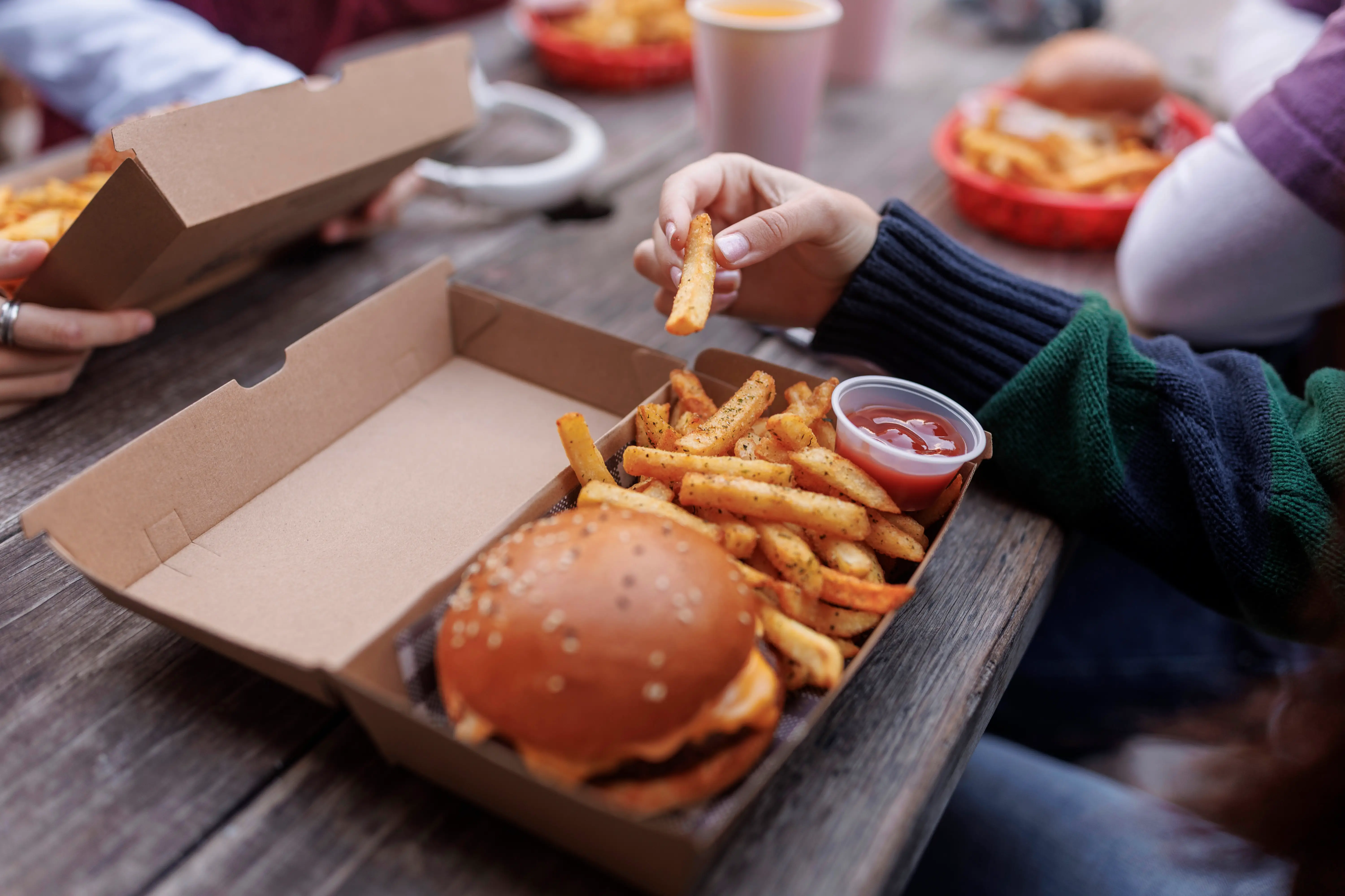 Hands reach for seasoned fries beside a burger and ketchup in a cardboard takeout box on a wooden table.