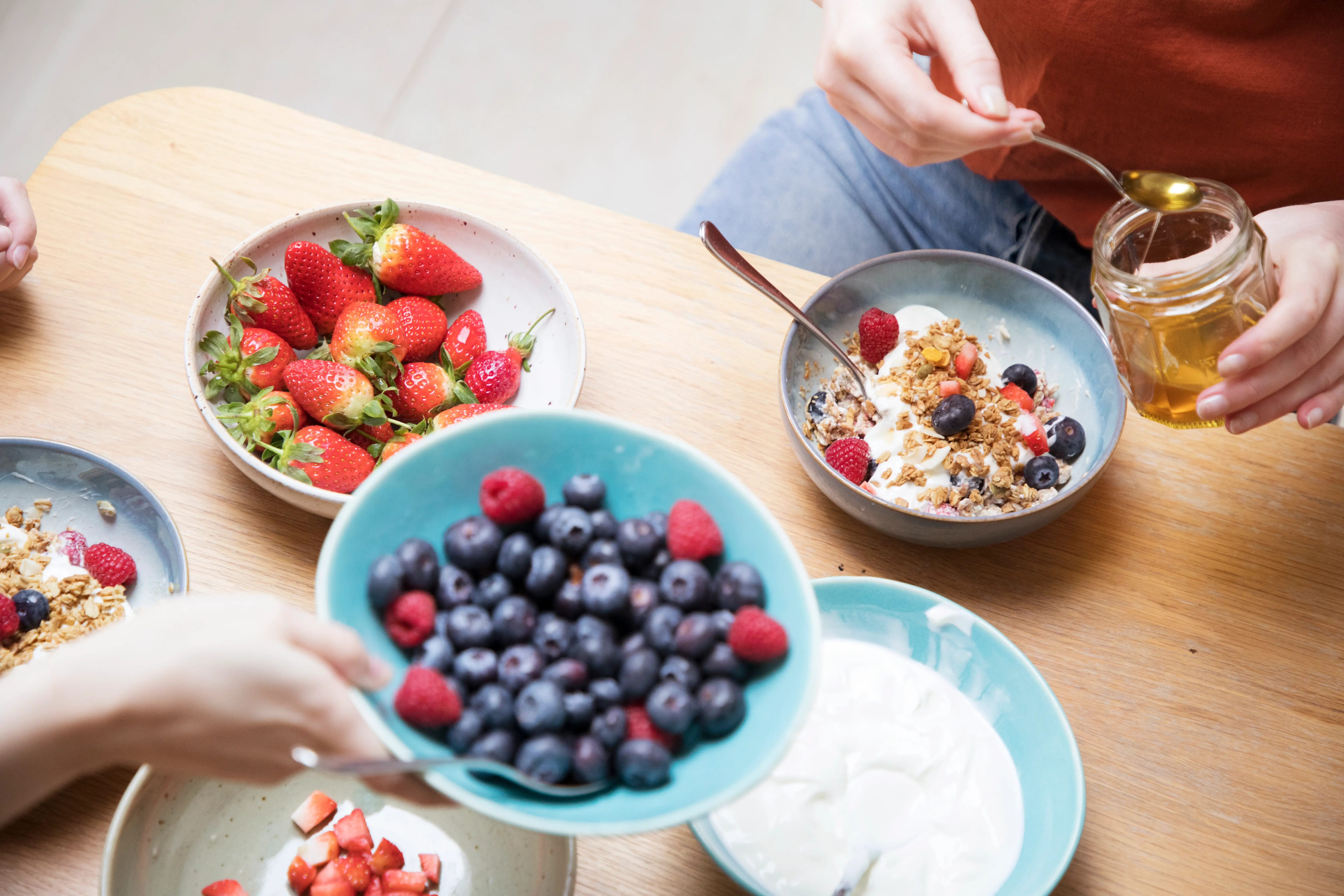 Hands prepare breakfast bowls with yogurt, granola, berries, strawberries, blueberries, raspberries, and a jar of honey on a wooden table.