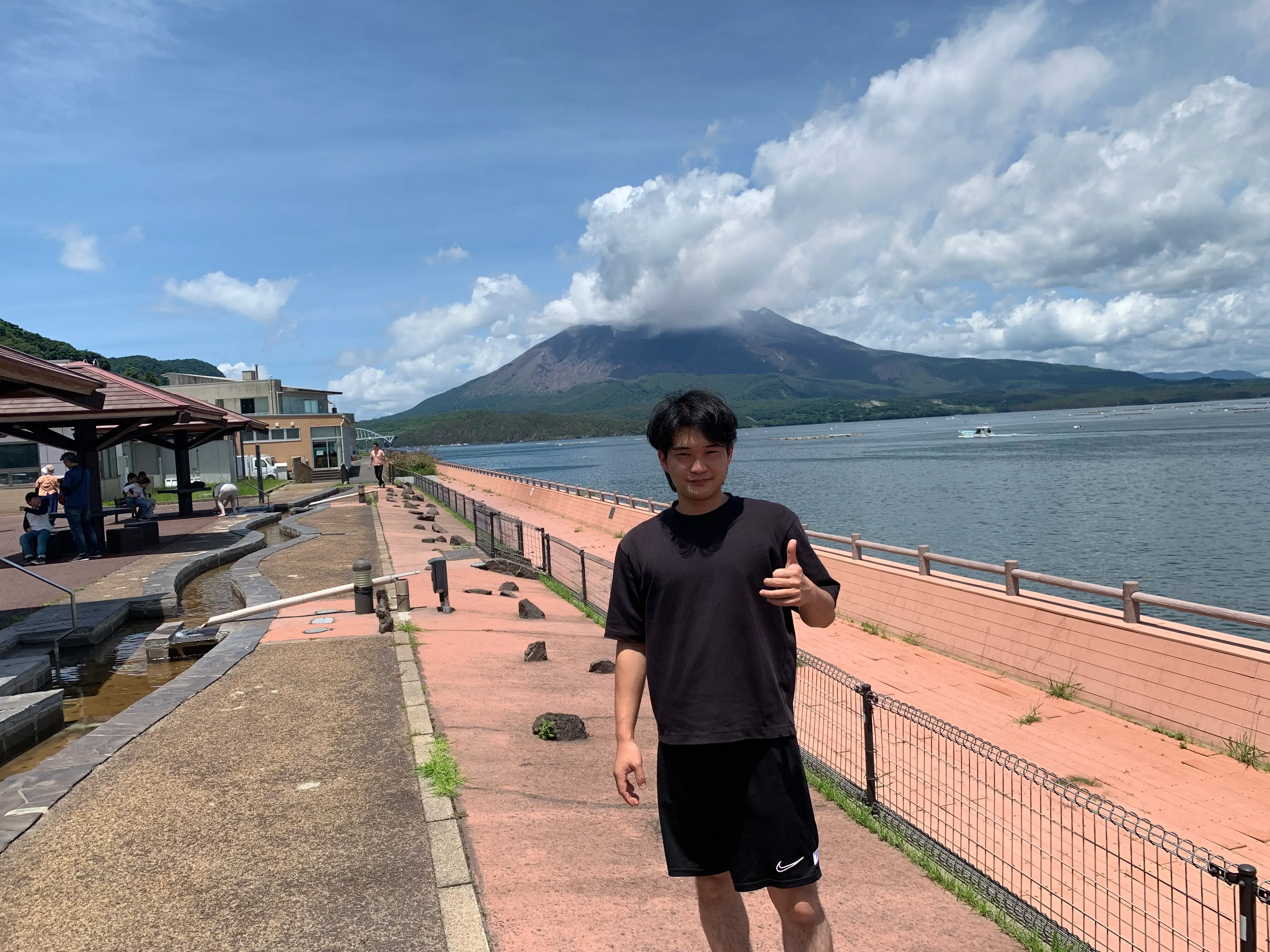 A man giving a thumbs up standing near the water with a mountain in Japan in the background.