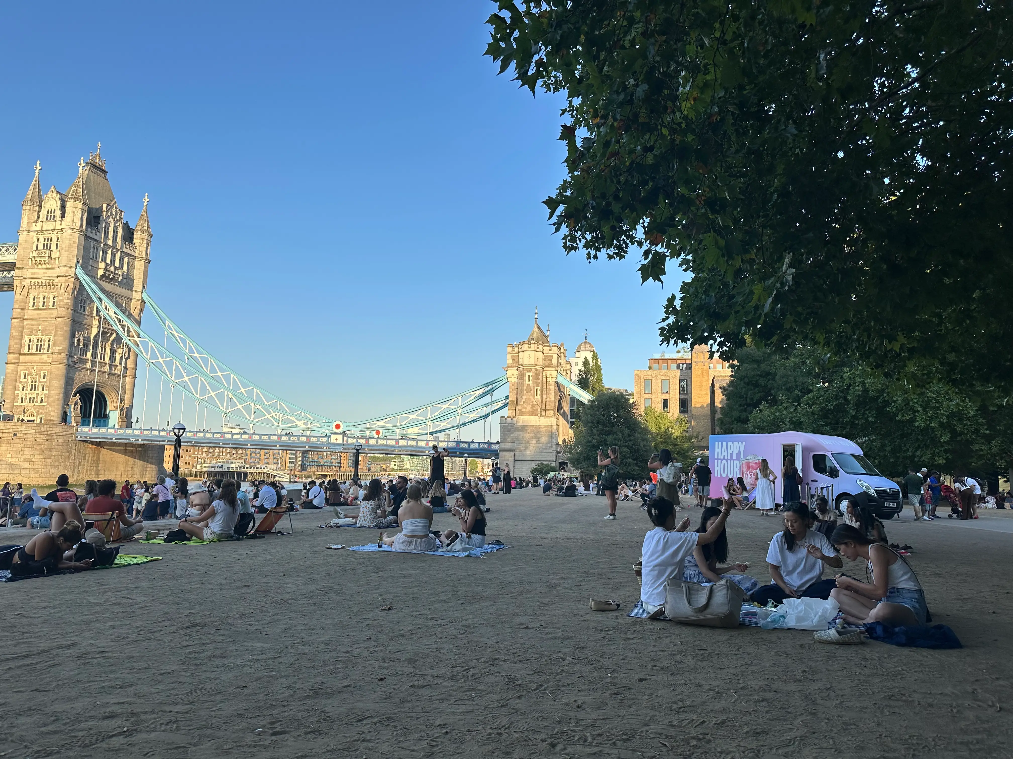 Crowds relax on a riverside lawn beside Tower Bridge in London under a clear blue sky.