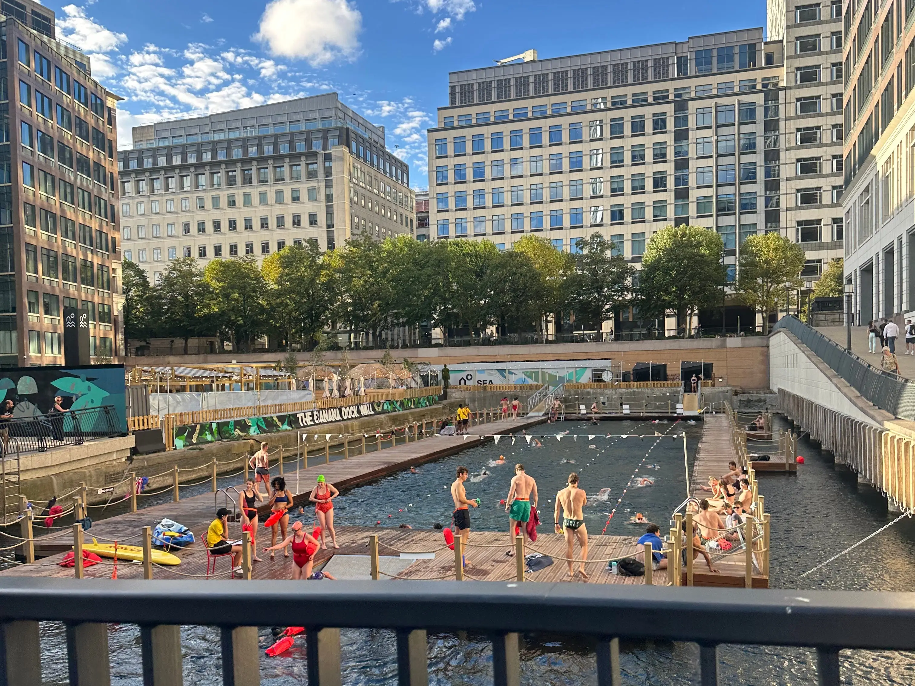 Swimmers gather on floating platforms at Sea Lanes, an urban dock pool surrounded by tall office buildings.