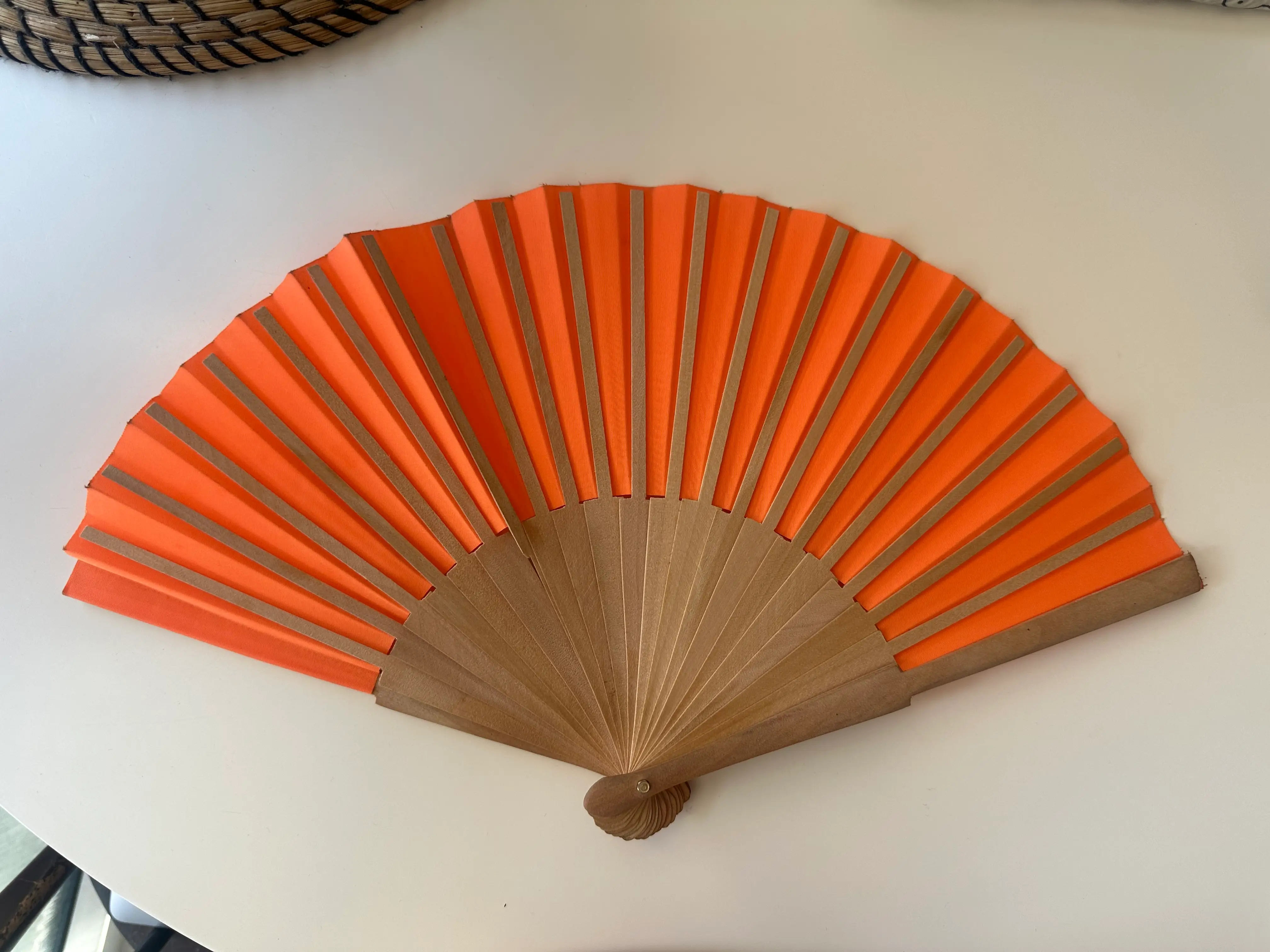 An open orange folding fan with wooden ribs rests on a light tabletop.