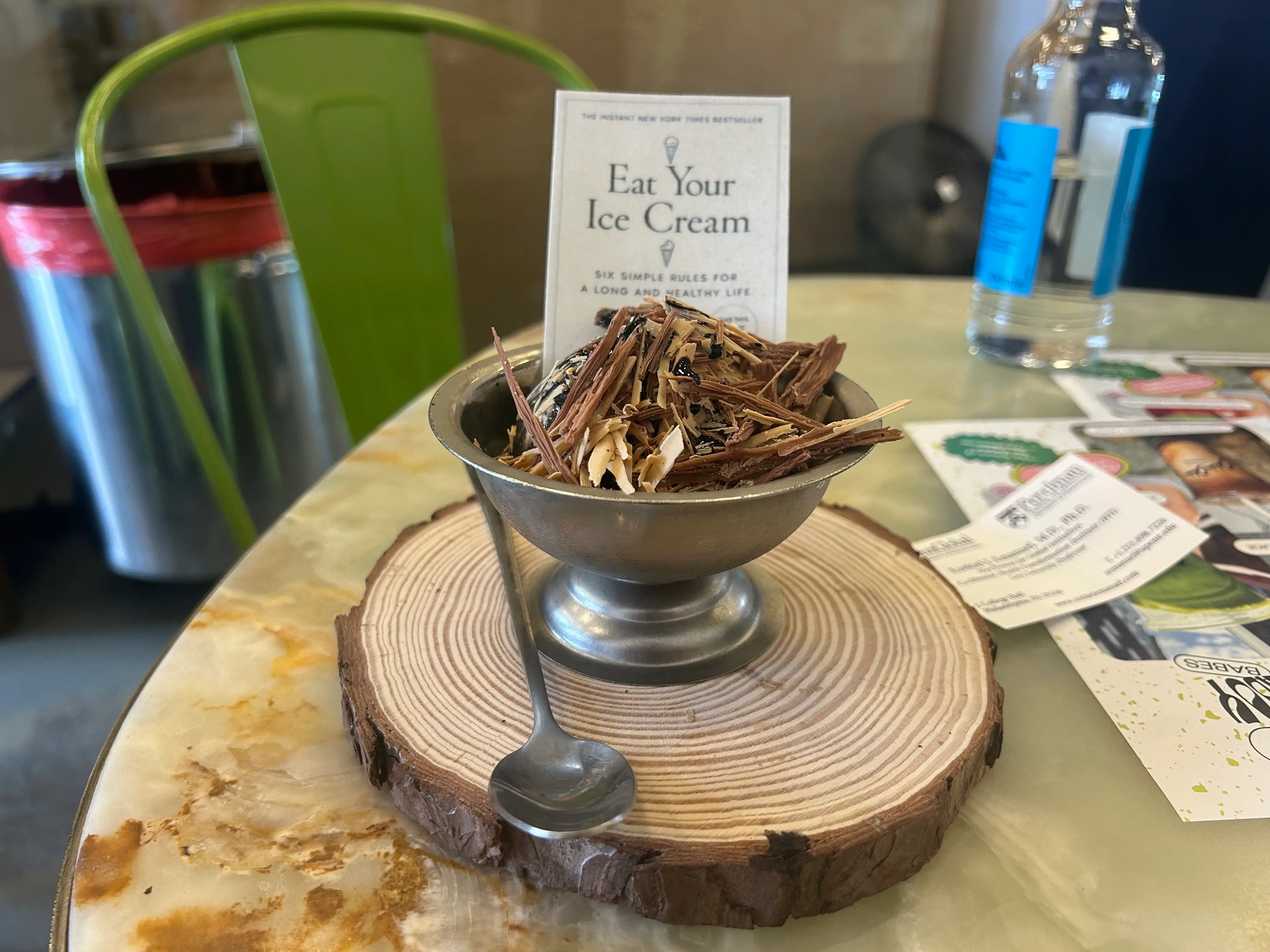 Metal dessert cup with shaved topping sits on a wood slice beside a spoon and “Eat Your Ice Cream” card.