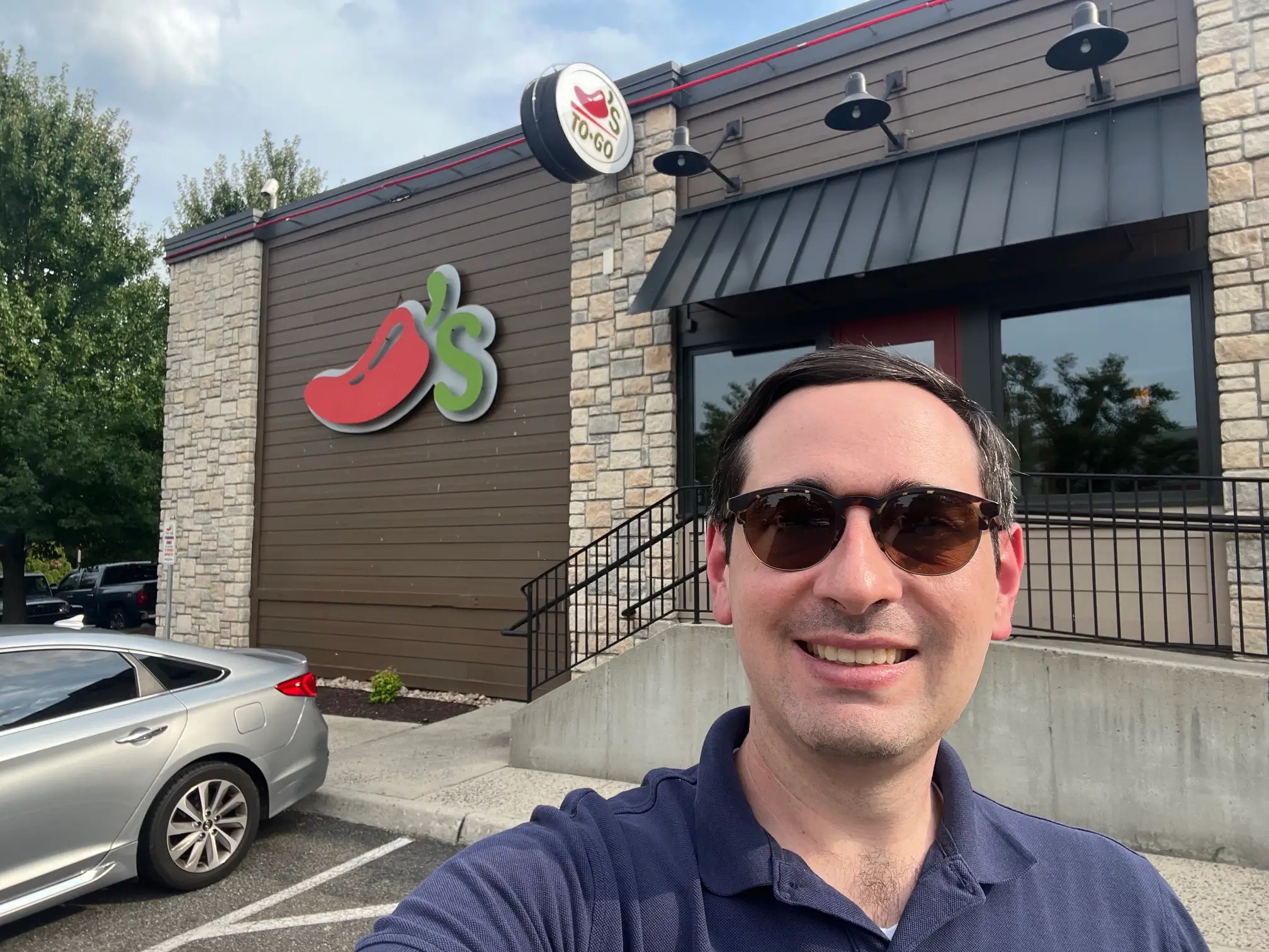 Person wearing sunglasses takes a selfie outside a Chili's restaurant with a TO GO sign visible.