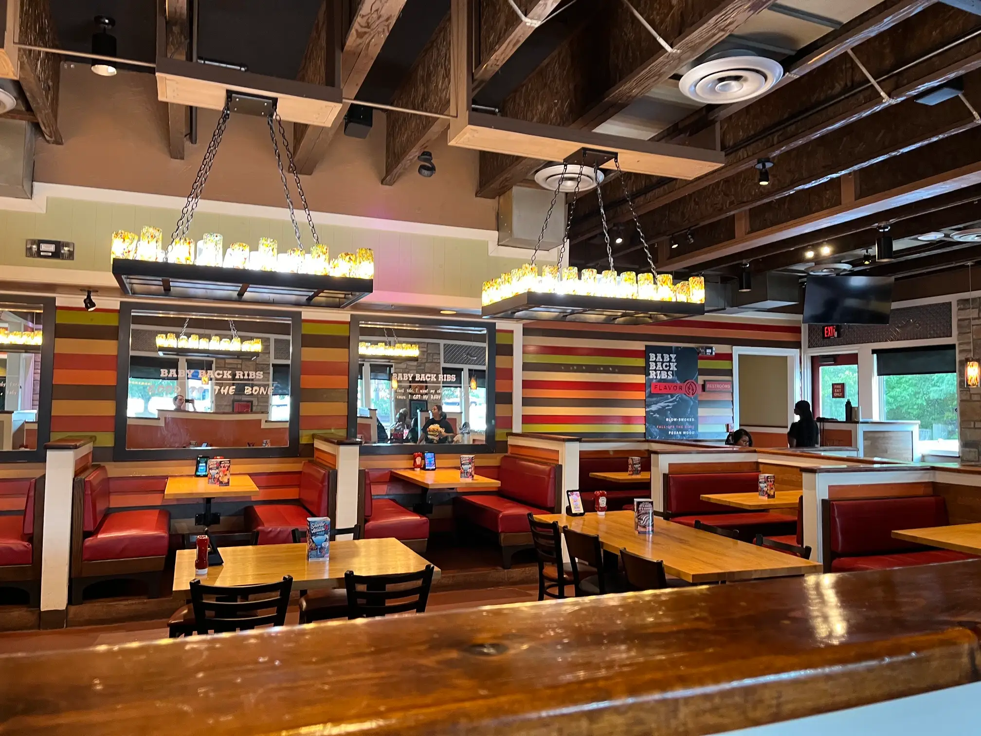 Empty restaurant dining area with red booths, wood tables, striped walls, hanging lights, and visible baby back ribs signage.