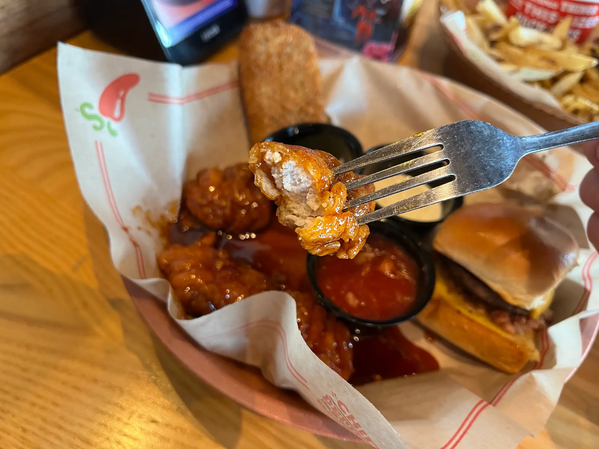 A fork holds a sauced chicken bite over a Chili’s basket with wings, burger, fries and dipping sauces.