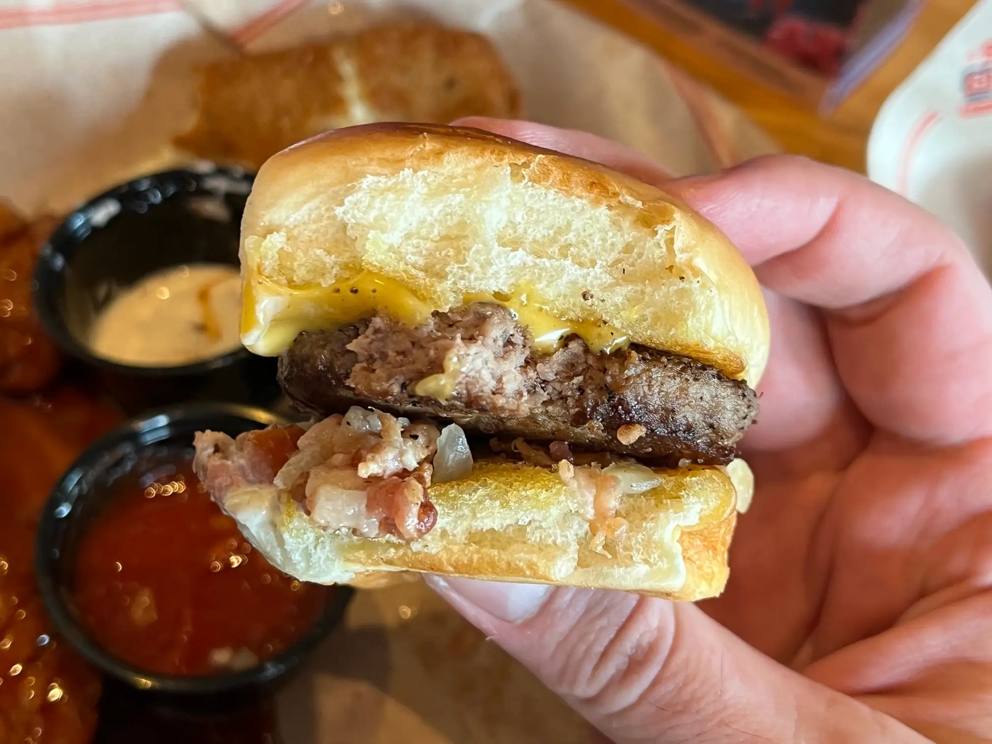Hand holding a small cheeseburger slider with melted cheese, bacon, onions, and dipping sauces nearby.