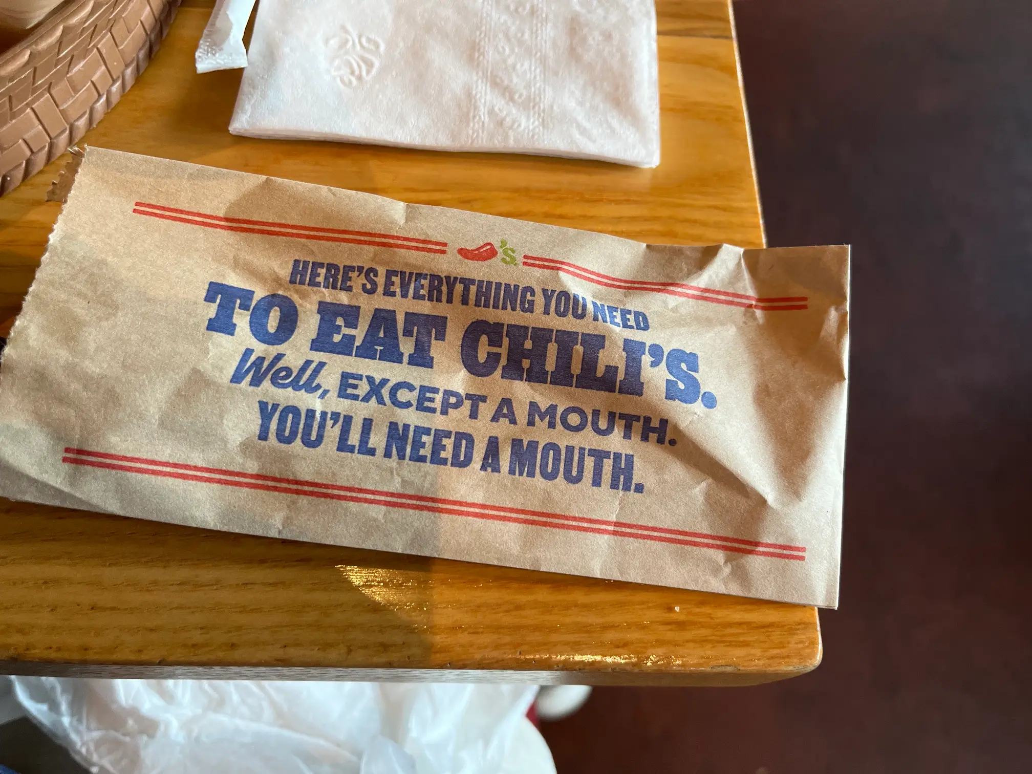 A Chili's paper sleeve with promotional text rests on a wooden table beside a folded napkin.