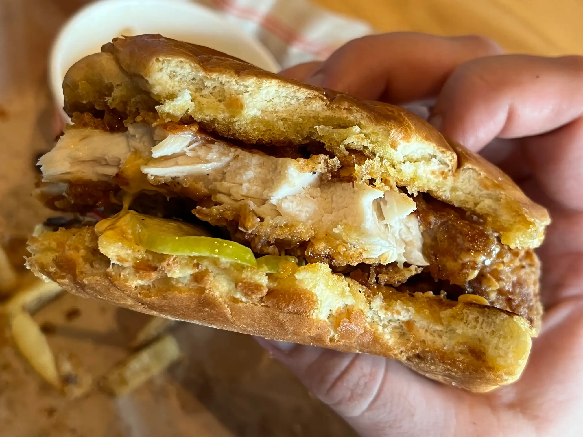 Close-up of a hand holding a partially eaten fried chicken sandwich with pickles and fries in the background.
