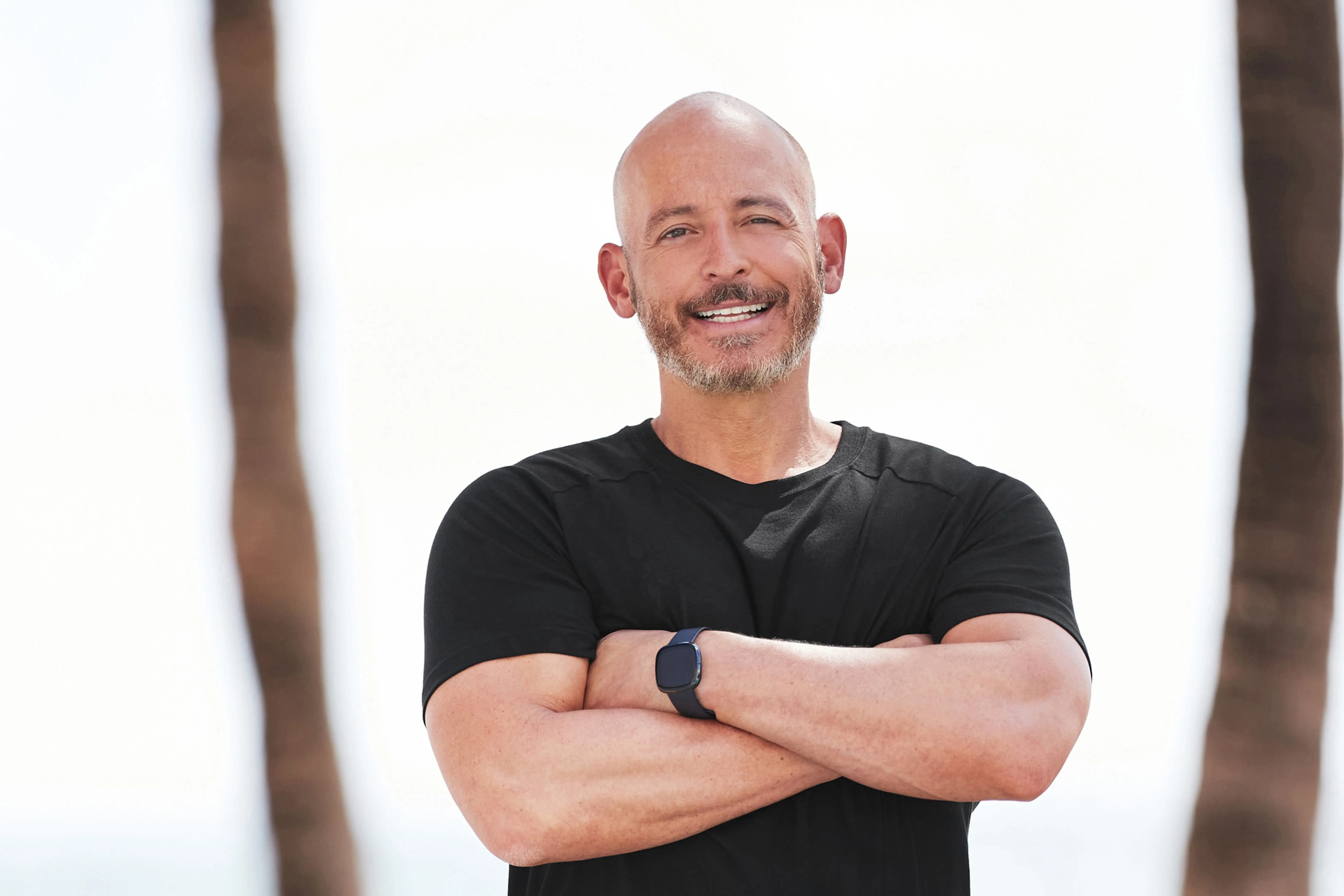 Smiling person in black athletic clothing stands with arms crossed near palm trees and a beach.