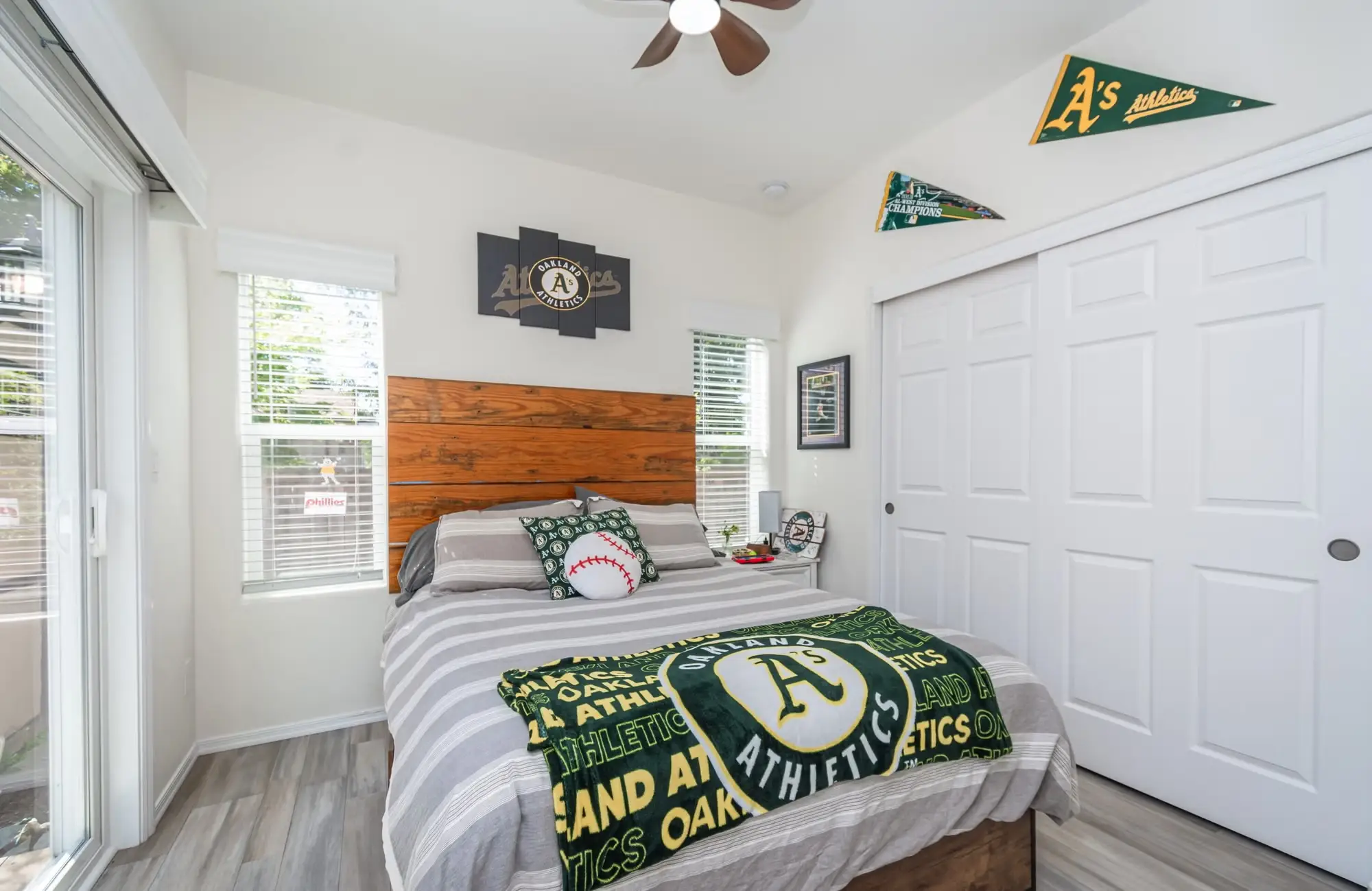 A bedroom with Oakland Athletics bedding, wall art, pennants, white closet doors, and a wooden headboard.