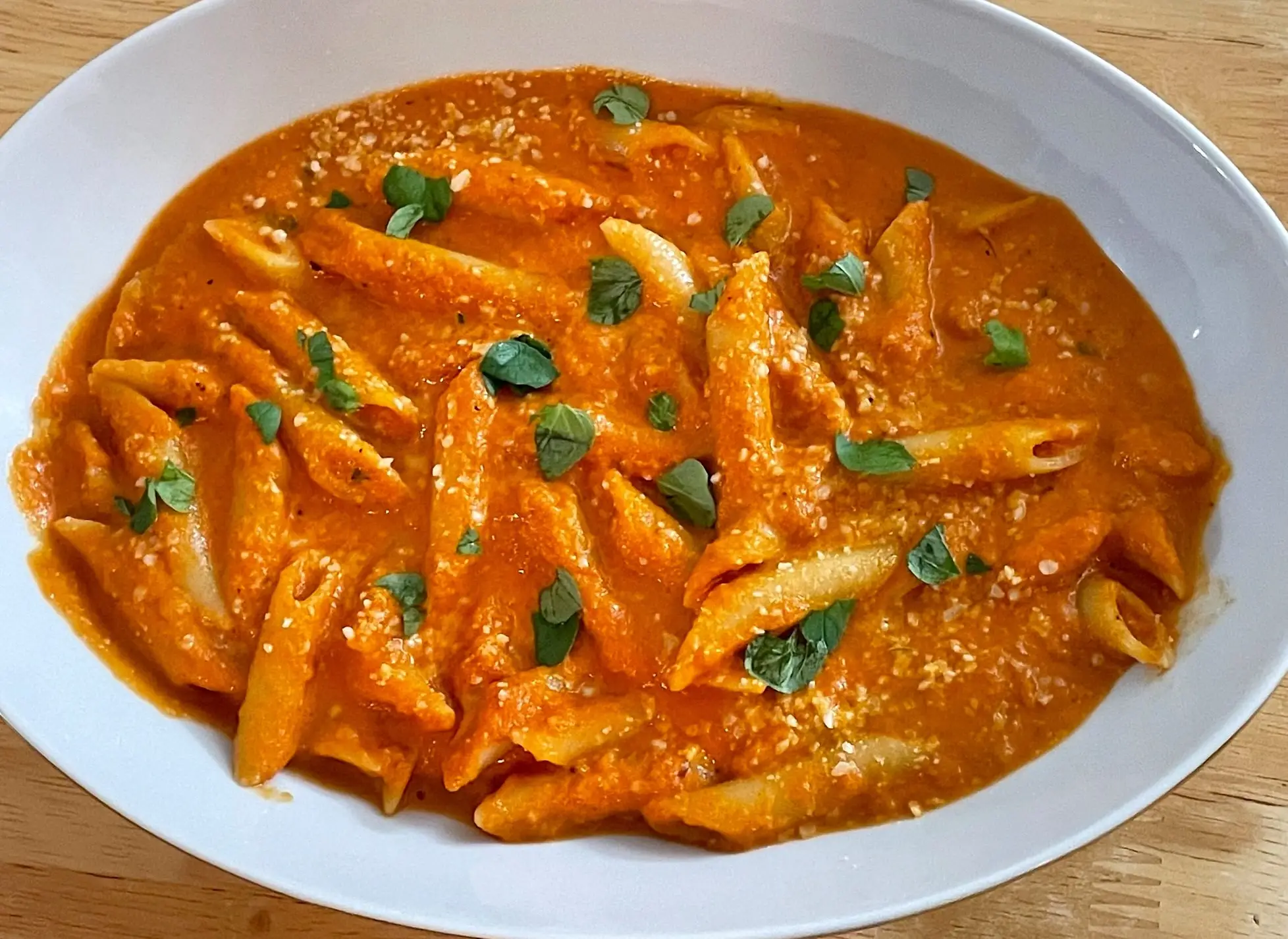 Penne pasta in orange tomato sauce topped with grated cheese and fresh green herbs on a white oval plate.