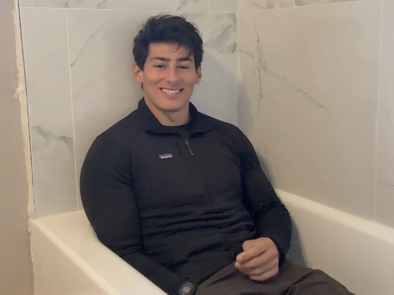 A smiling person in a black Patagonia pullover sits inside a white bathtub with marble-patterned tile walls.
