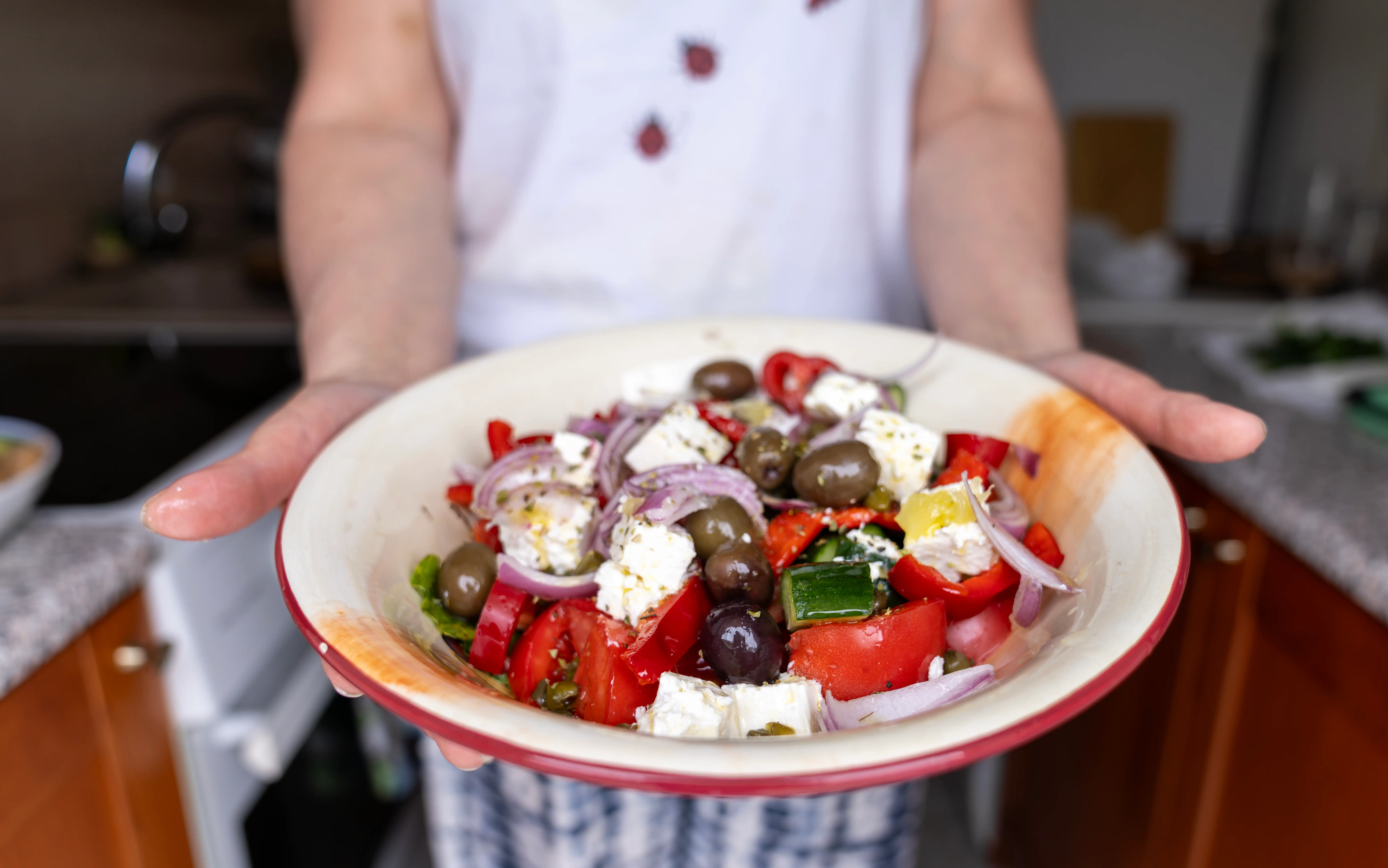 A person serving a plate of food that includes red pepper, red onion, olives, cucumber, and feta.